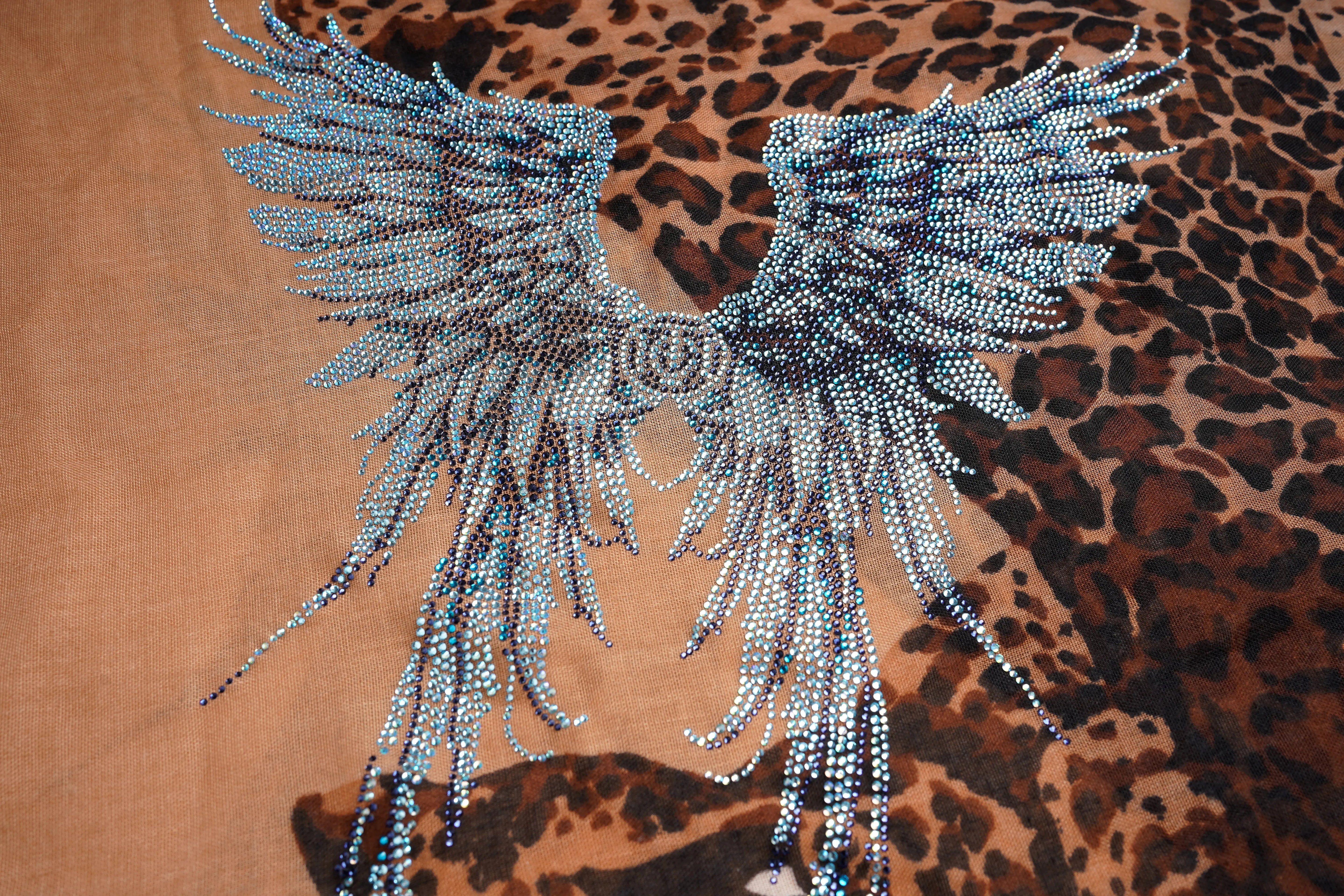 A close-up of a leopard-print fabric featuring a large embroidered angel wing design in light blue and white beads with a central rose motif. TCH Rhinestone Winged Leopard Lightweight Fashion Scarf