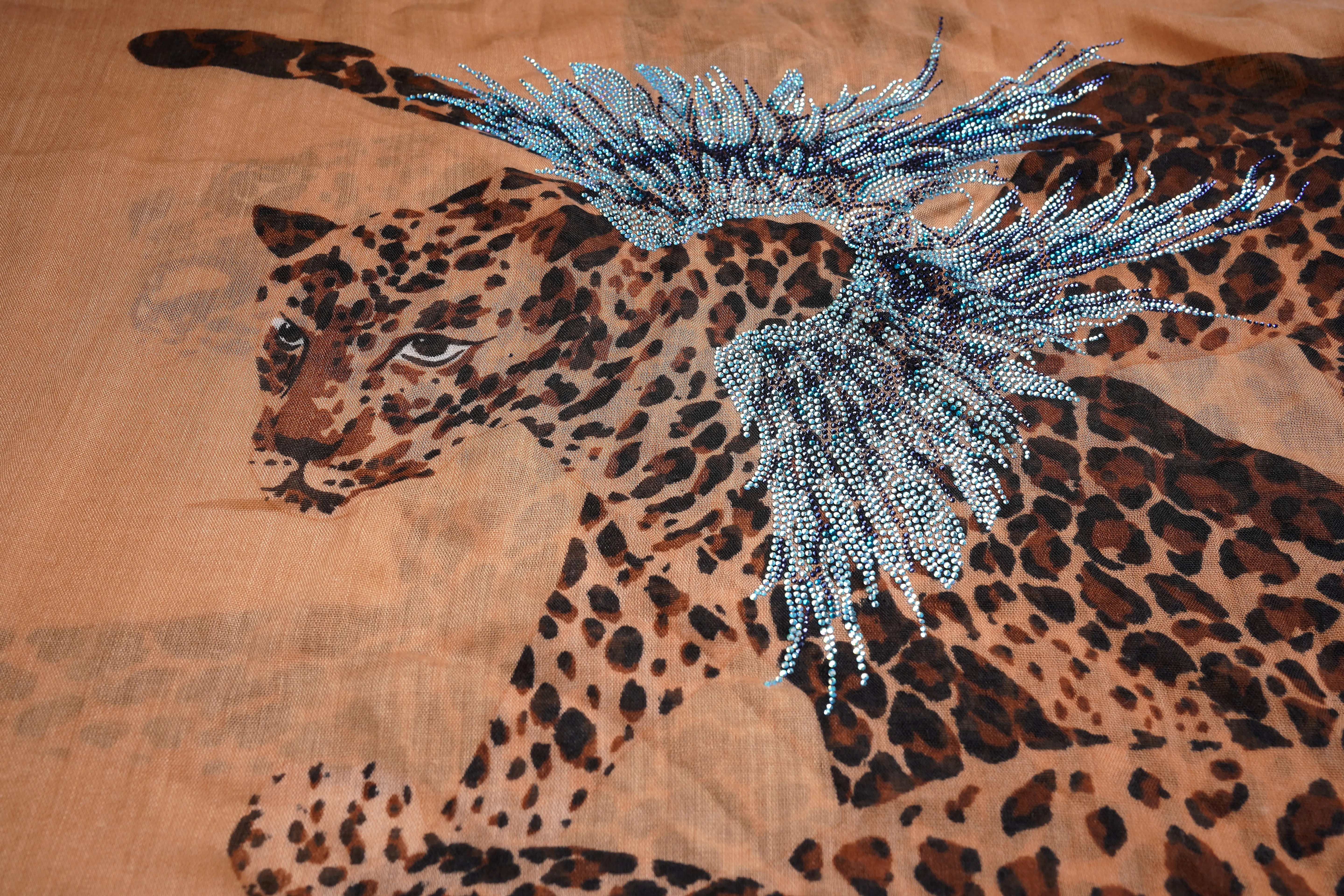 A close-up of a leopard print fabric featuring a detailed illustration of a leopard in brown and black tones, overlaid with a decorative blue beaded feather-like embellishment. TCH Rhinestone Winged Leopard Lightweight Fashion Scarf