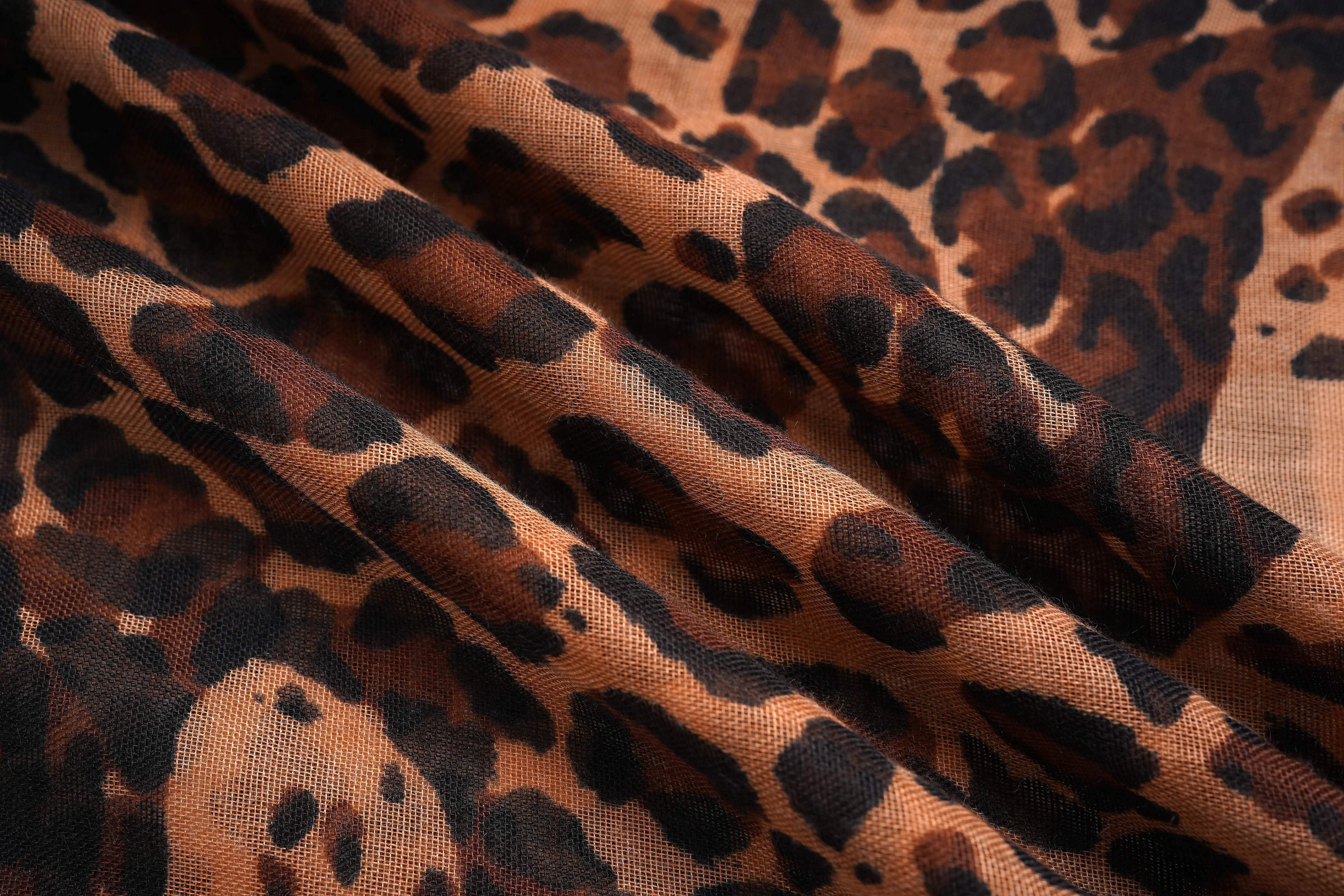A close-up of leopard print fabric with a warm tan base and dark brown to black irregular spots, shown in soft folds. TCH Rhinestone Winged Leopard Lightweight Fashion Scarf
