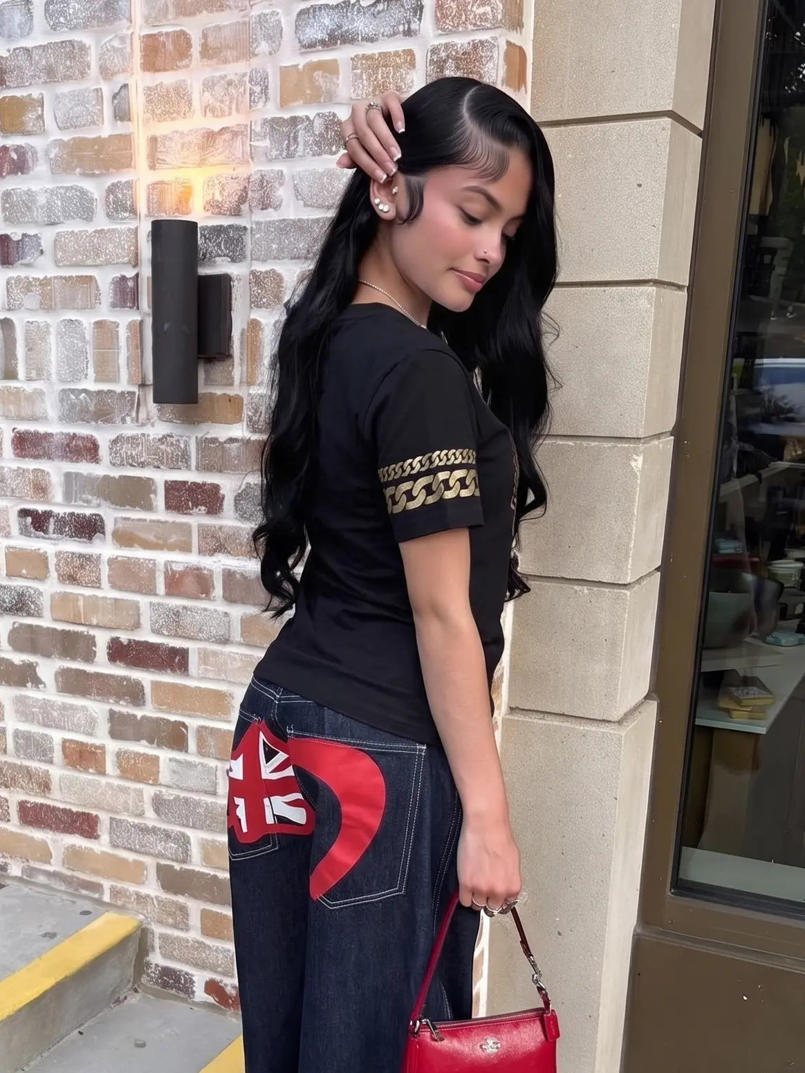 Woman wearing a black t-shirt with gold chain design and dark jeans with red heart design, standing against a brick wall. TCH Rhinestone Winged Unicorn T-Shirt