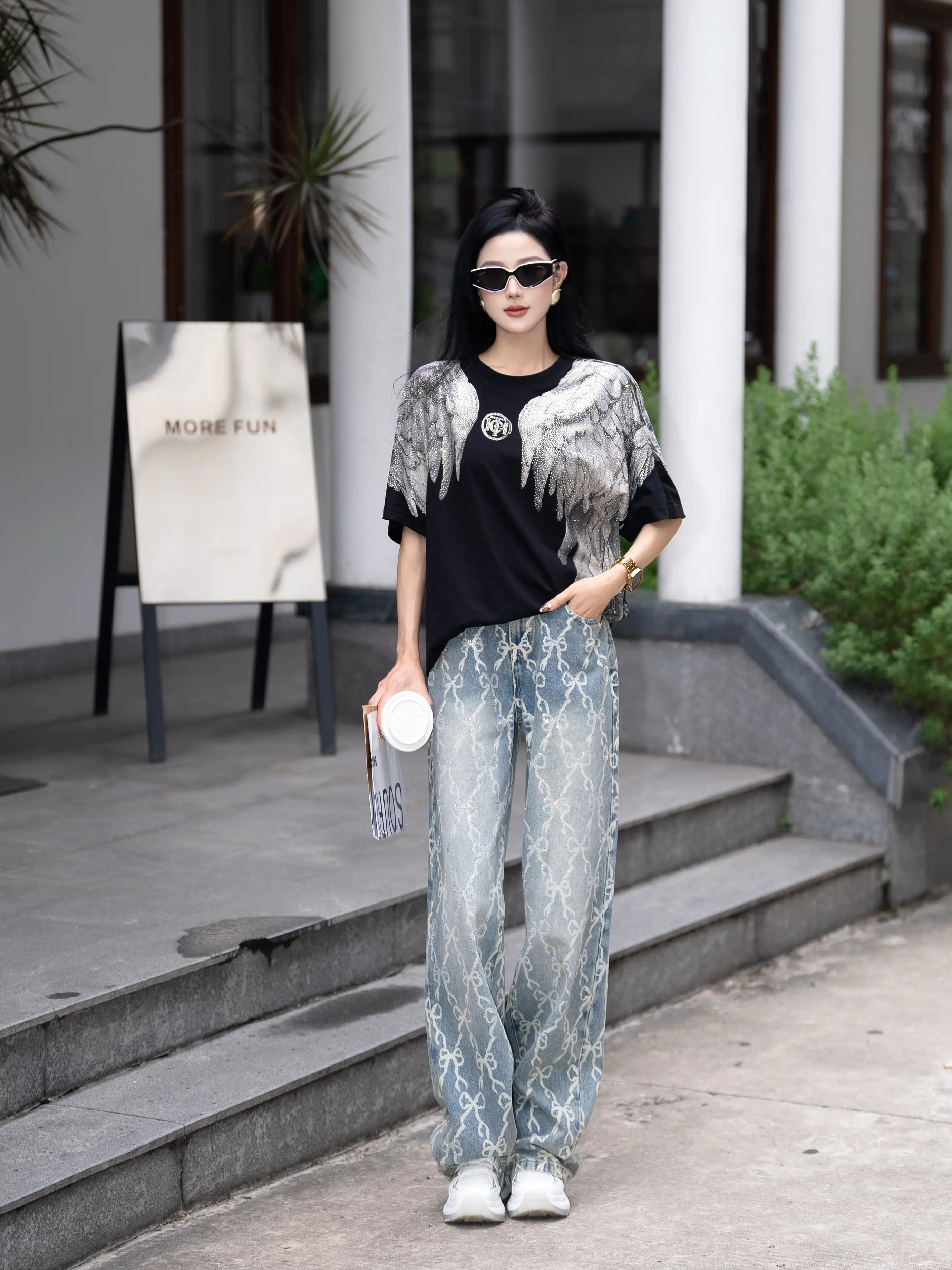 Woman in stylish outfit standing outdoors with a building and plants in the background, TCH Rhinestone Wings Logo Black Graphic T-Shirt