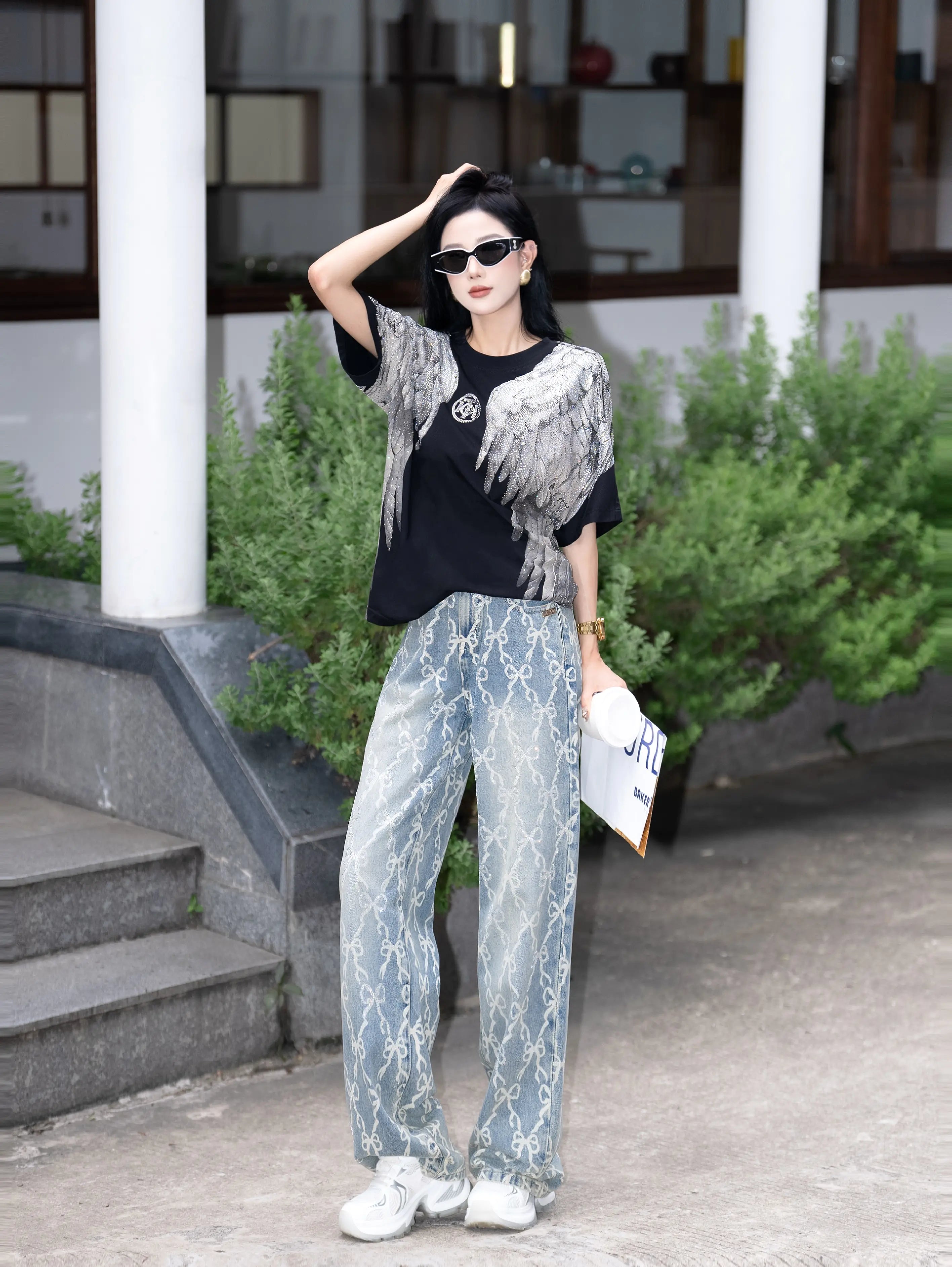 Woman in stylish outfit standing outdoors with greenery and a building in the background, TCH Rhinestone Wings Logo Black Graphic T-Shirt