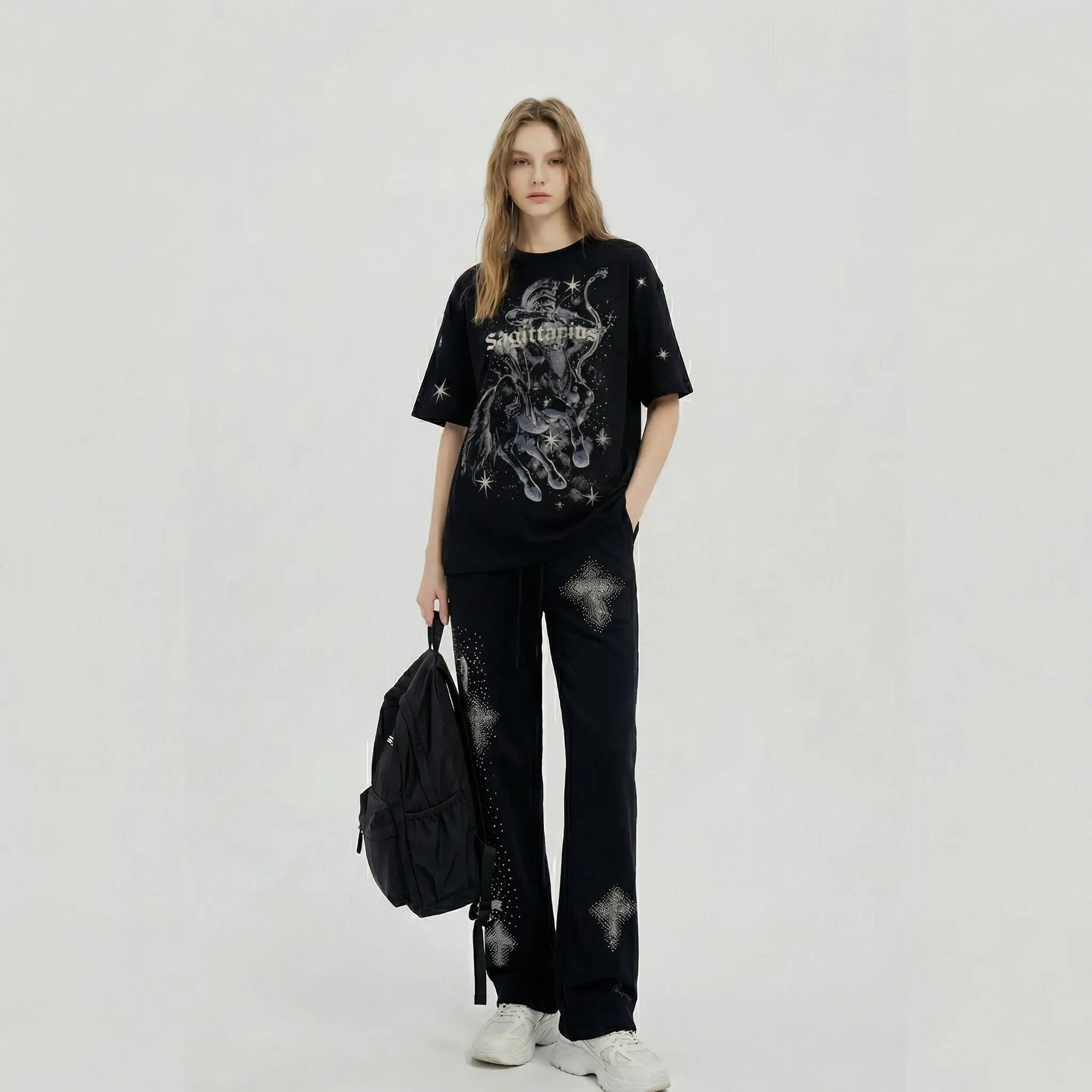 A model stands against a light gray studio background wearing a black graphic T-shirt with a white gothic-style illustration and text, matching black pants with distressed and cross motifs, white sneakers, and carrying a black backpack. TCH Sagittarius Celestial Graphic Oversized T-Shirt