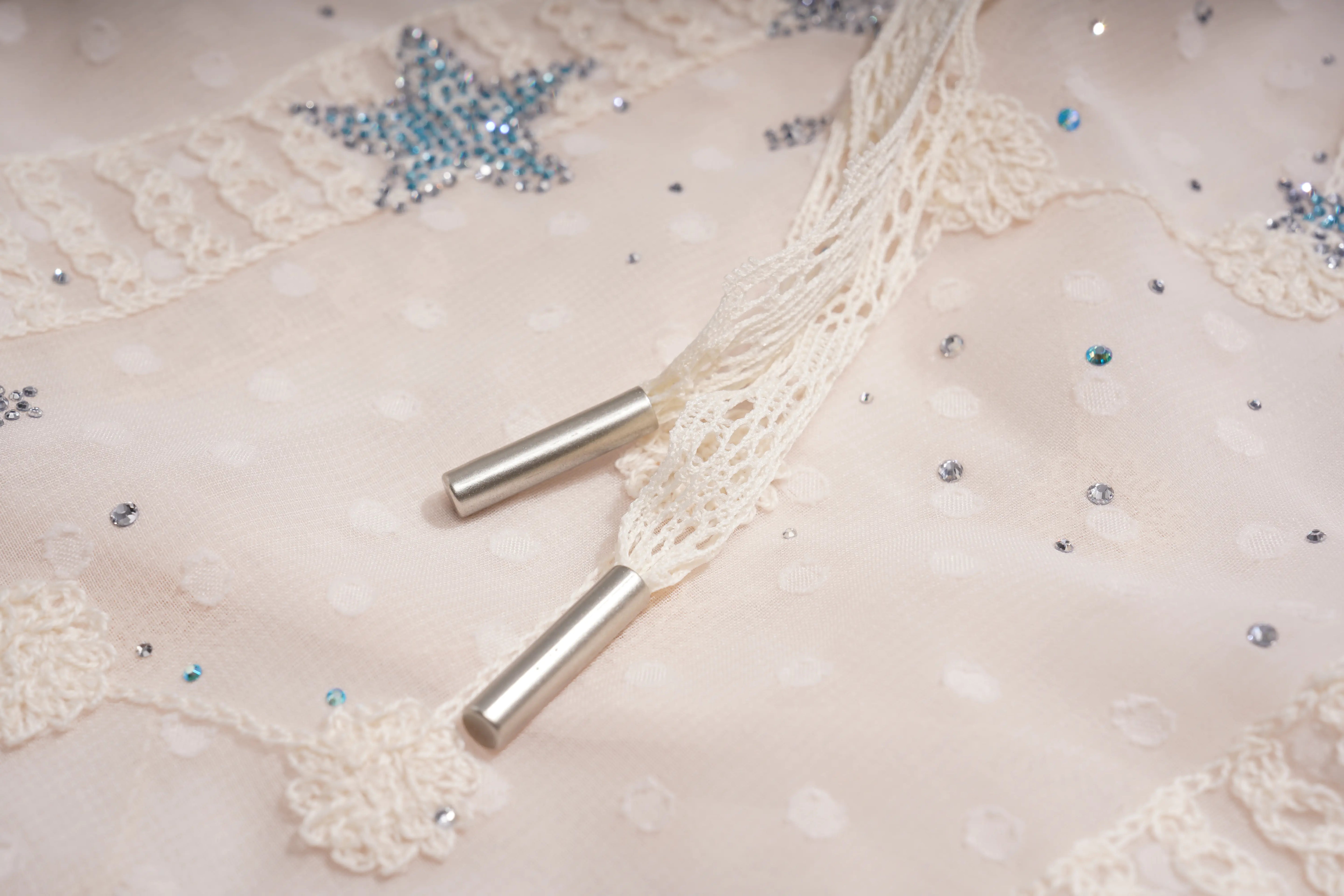 Close-up of a cream lace drawstring with silver cylindrical metal tips on a light beige embellished fabric with sequins and rhinestones. TCH Sheer Rhinestone Angel Star Zip-Up Hoodie