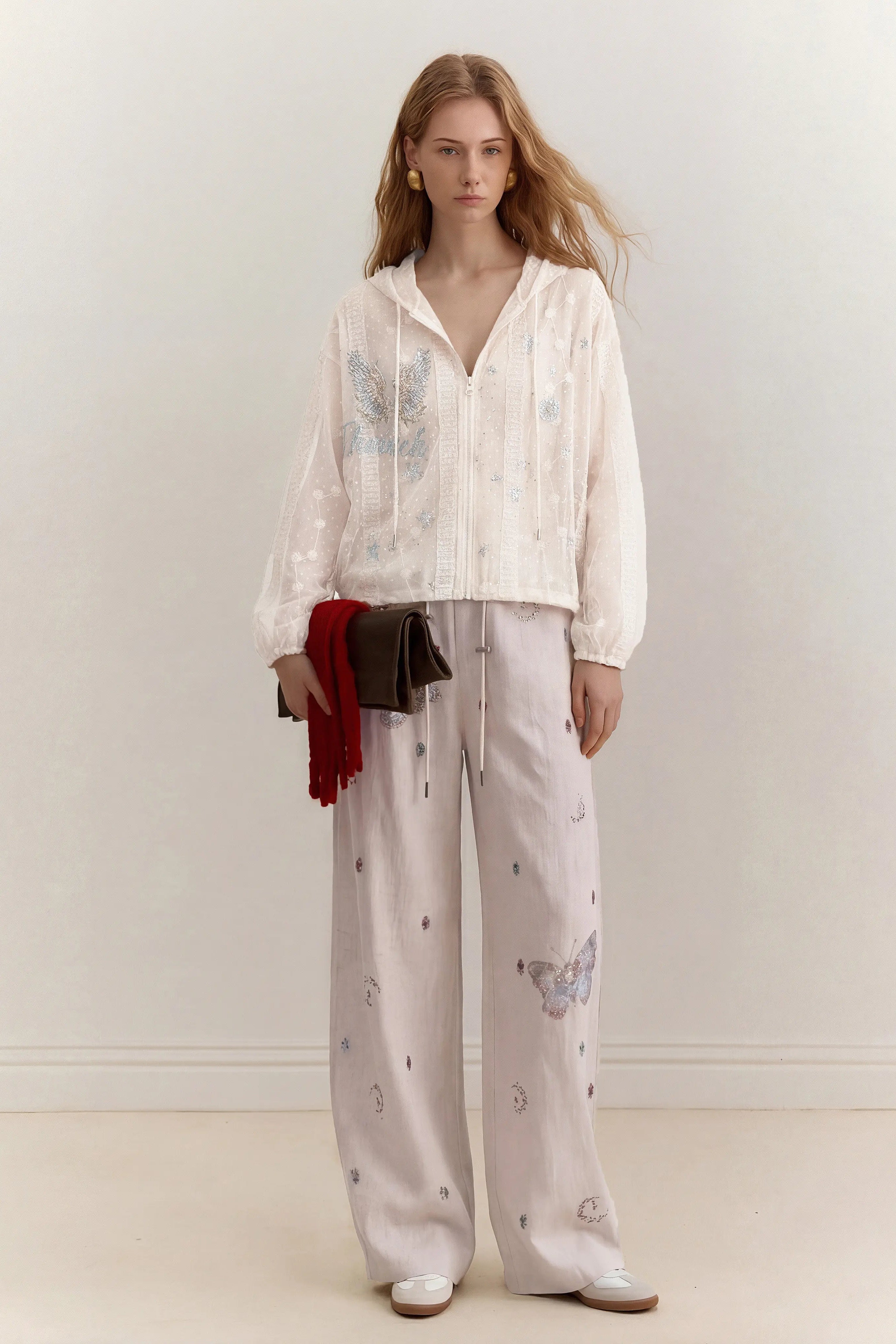 A woman stands in a minimalist studio setting wearing a white embroidered long-sleeve top and matching wide-leg trousers with butterfly and moon motifs. She holds a small dark clutch with a red fringed scarf draped over it. TCH Sheer Rhinestone Angel Star Zip-Up Hoodie