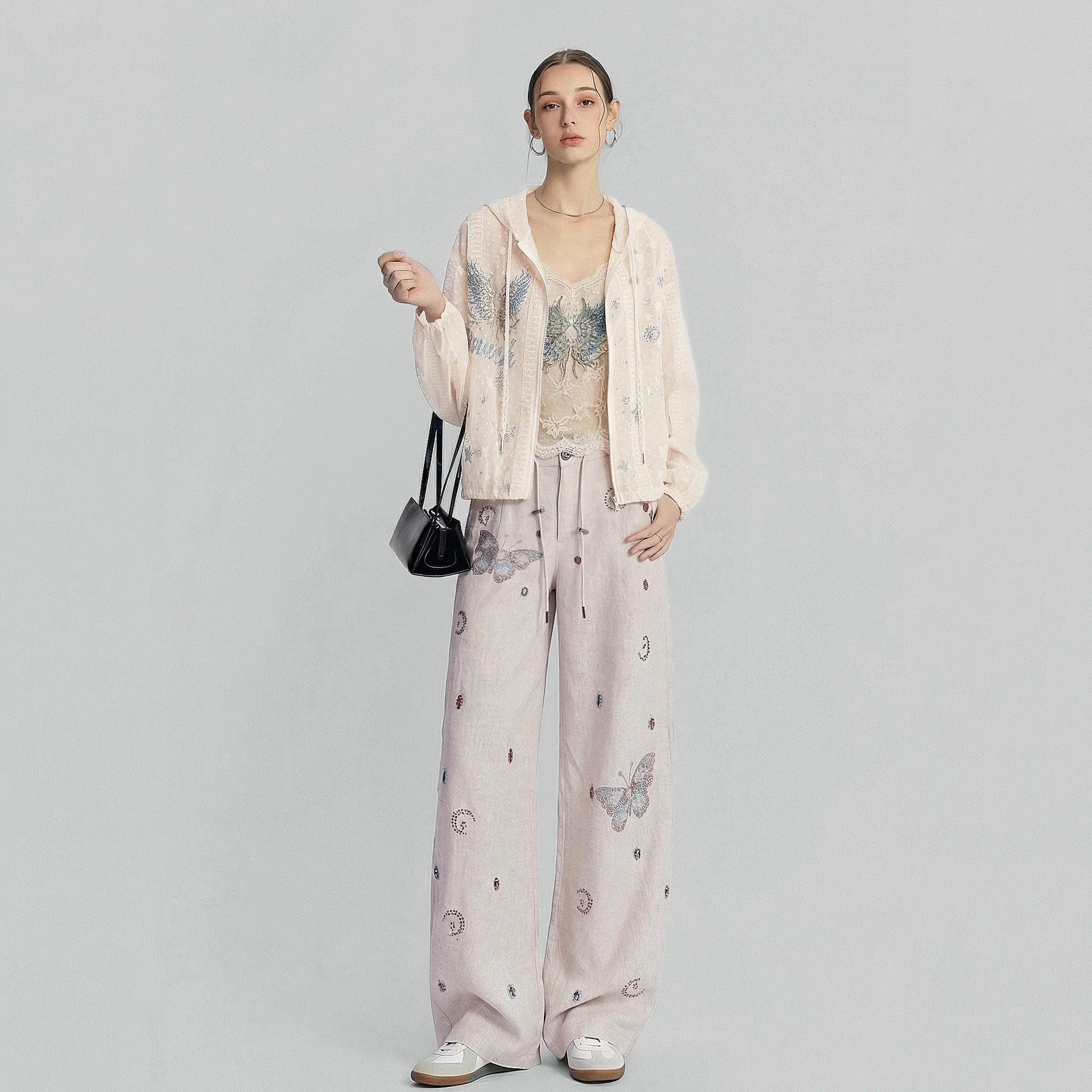A model stands against a light gray studio background wearing a matching light pink lounge set consisting of a long-sleeve open-front cardigan and wide-leg pants with butterfly and moon prints. She is also wearing a beige lace-trimmed top, white sneakers, and carrying a small black handbag. TCH Sheer Rhinestone Angel Star Zip-Up Hoodie
