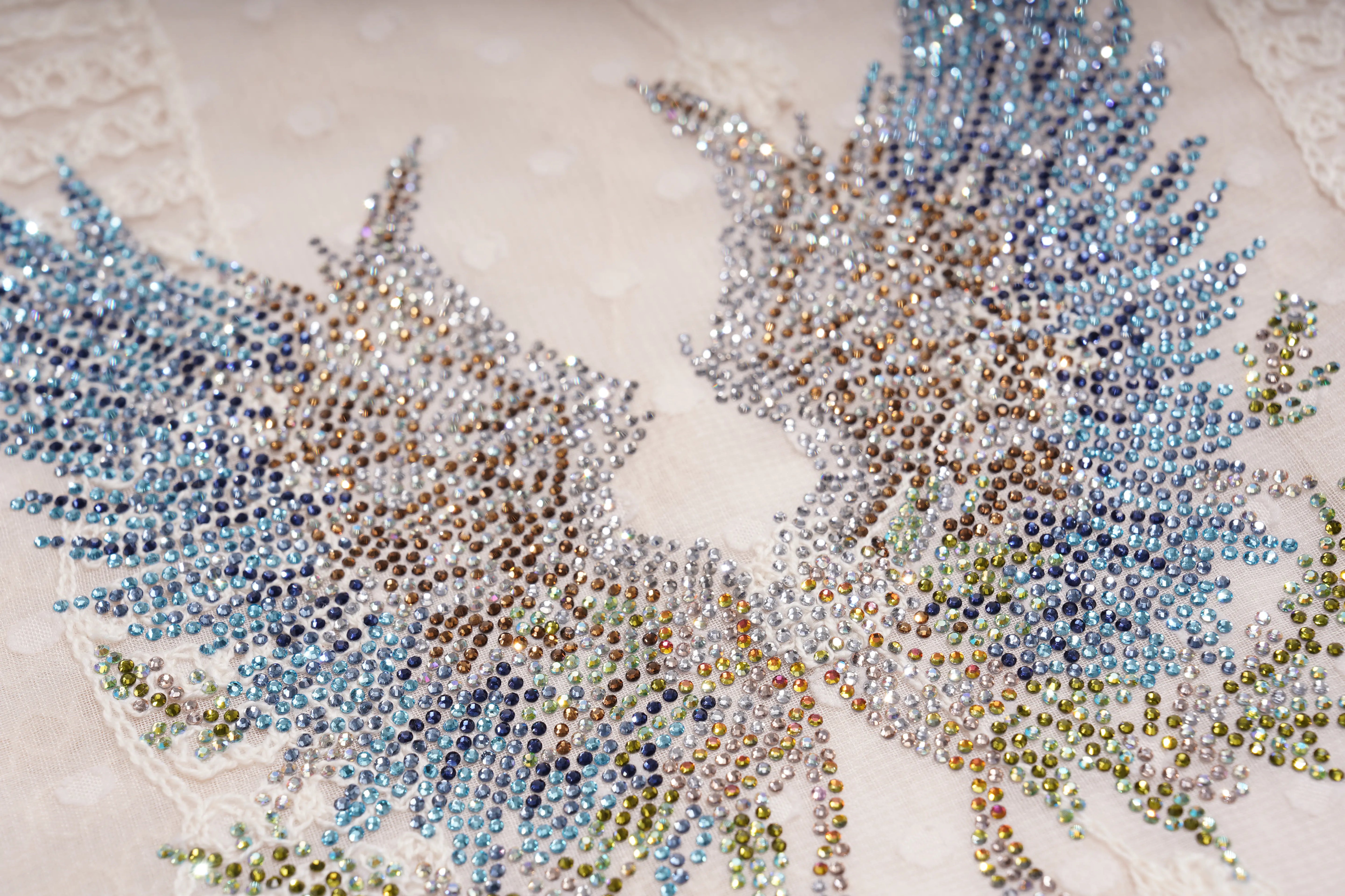 Close-up of a decorative embroidered or beaded wing motif on a light cream fabric, featuring iridescent rhinestones that shift between silver, blue, gold, and green. TCH Sheer Rhinestone Angel Star Zip-Up Hoodie