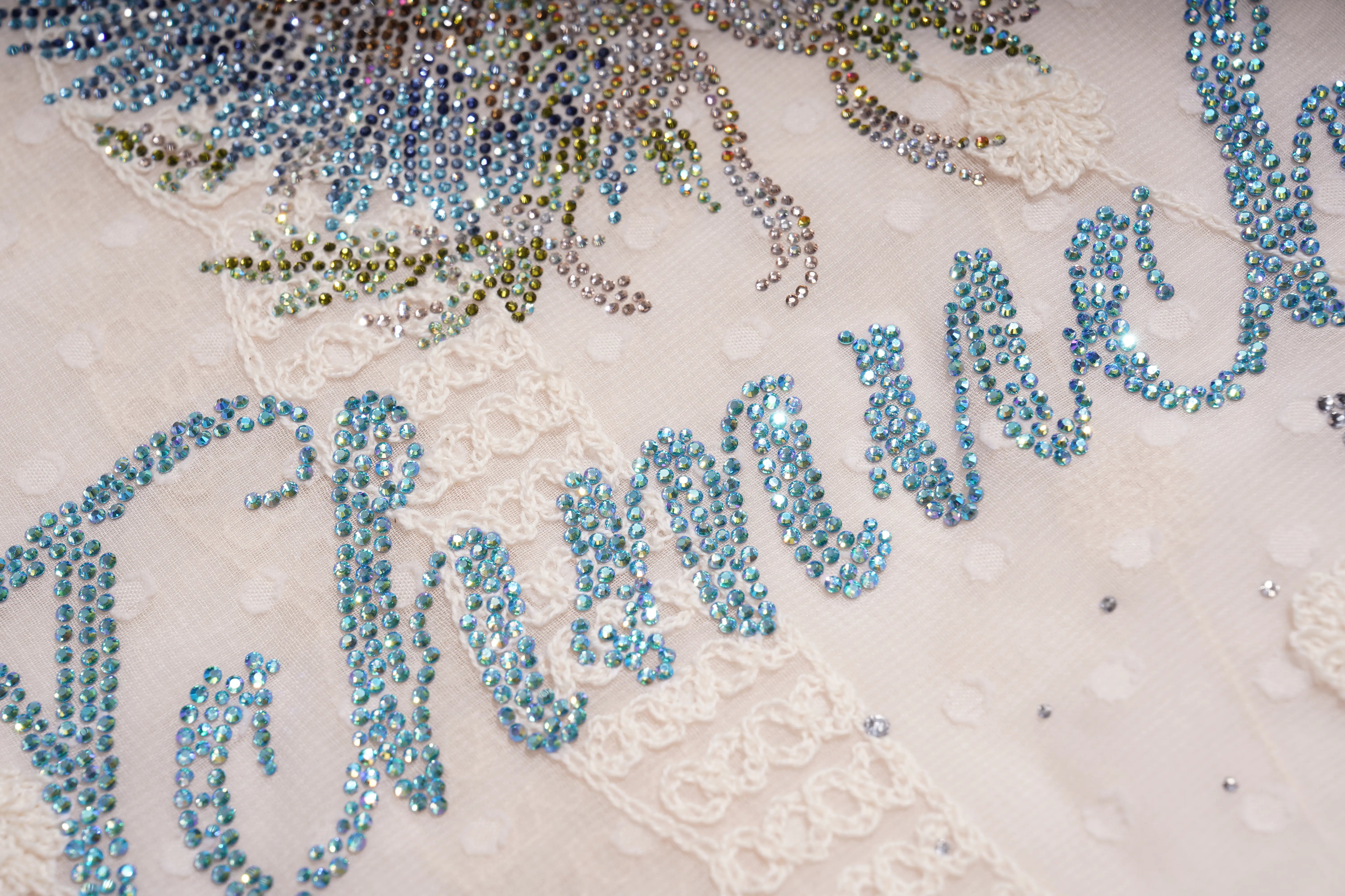 Close-up of a cream lace garment embellished with light blue rhinestone lettering and a floral design in blue, green, and silver beads. TCH Sheer Rhinestone Angel Star Zip-Up Hoodie