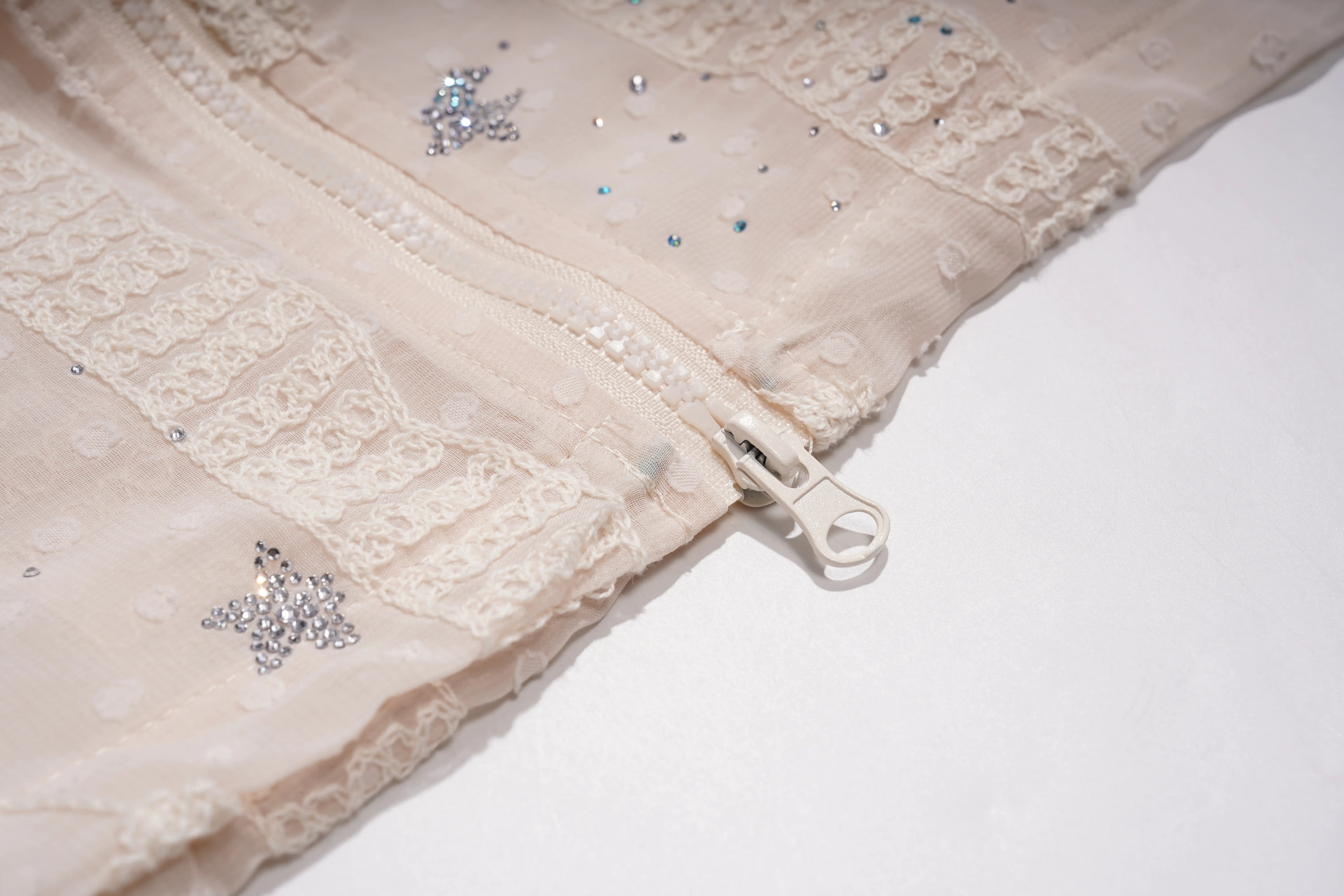 Close-up of a cream-colored lace garment with rhinestone star embellishments and a silver zipper pull, shown on a white background. TCH Sheer Rhinestone Angel Star Zip-Up Hoodie
