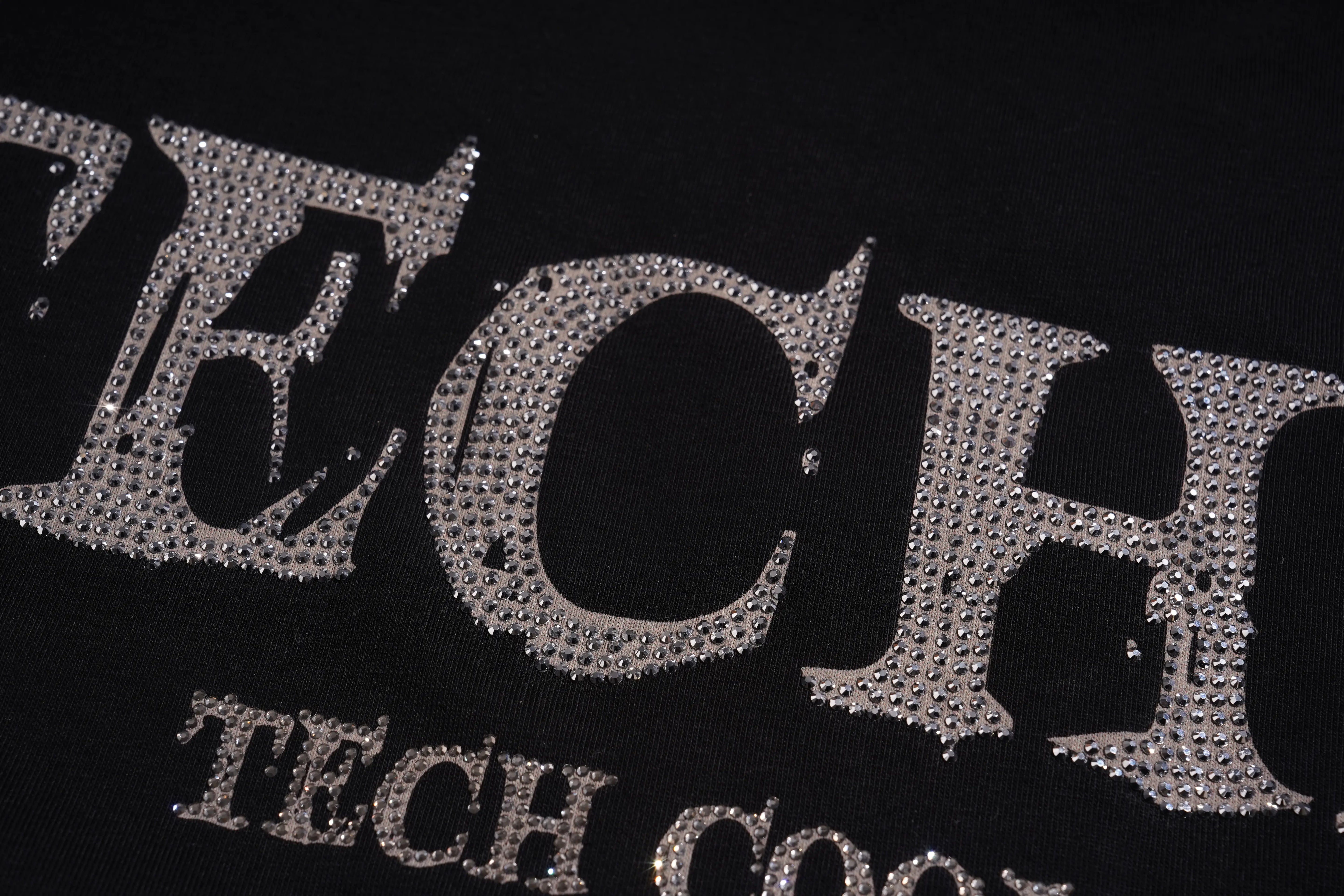 Close-up of a black garment featuring large silver rhinestone-embellished letters spelling part of a graphic, with smaller matching text below. TCH Silver Wing Techno Oversized Graphic T-Shirt