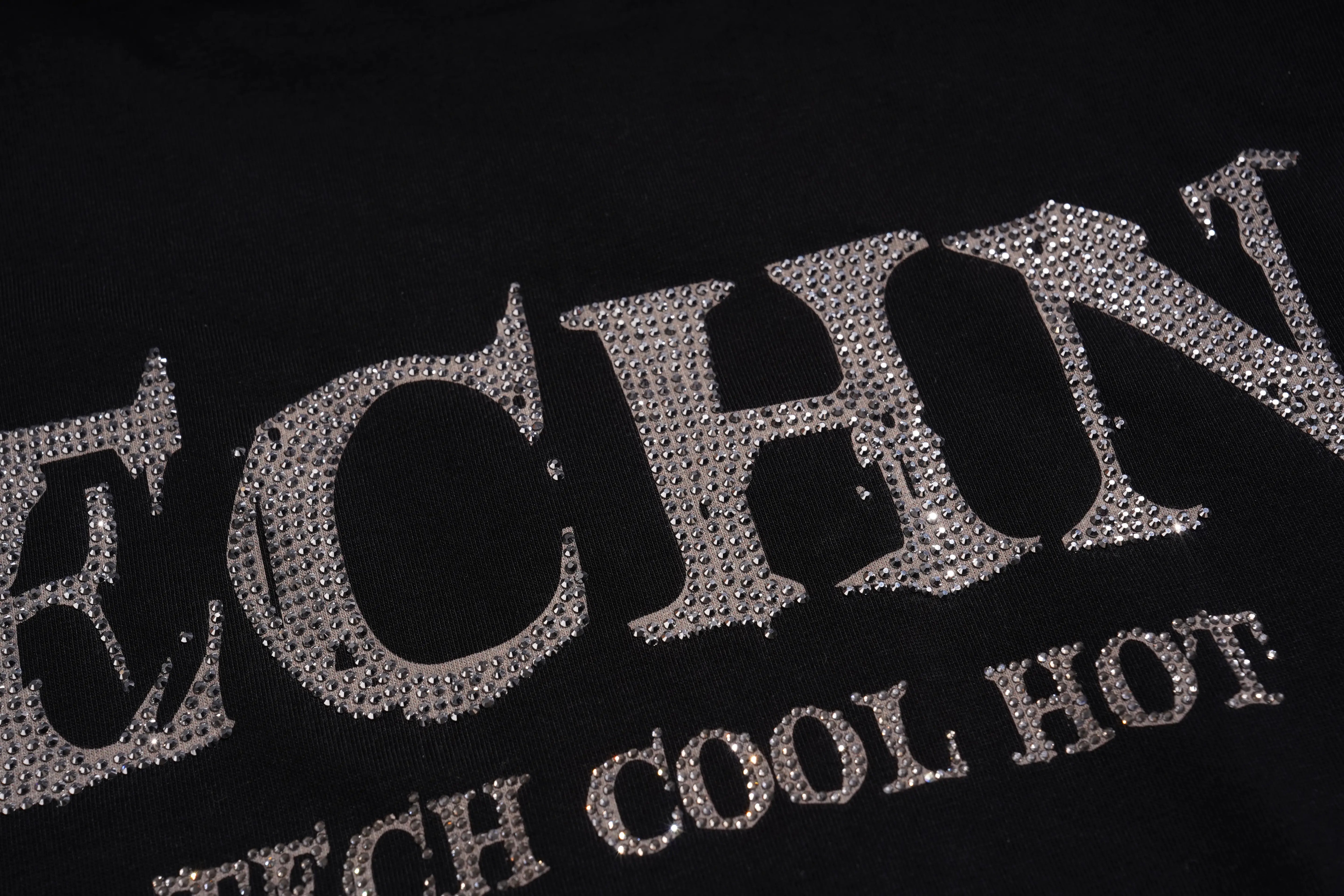 A close-up of a black garment featuring large, silver rhinestone-embellished letters spelling 'TECH' and smaller text reading 'COOL HOT'. TCH Silver Wing Techno Oversized Graphic T-Shirt