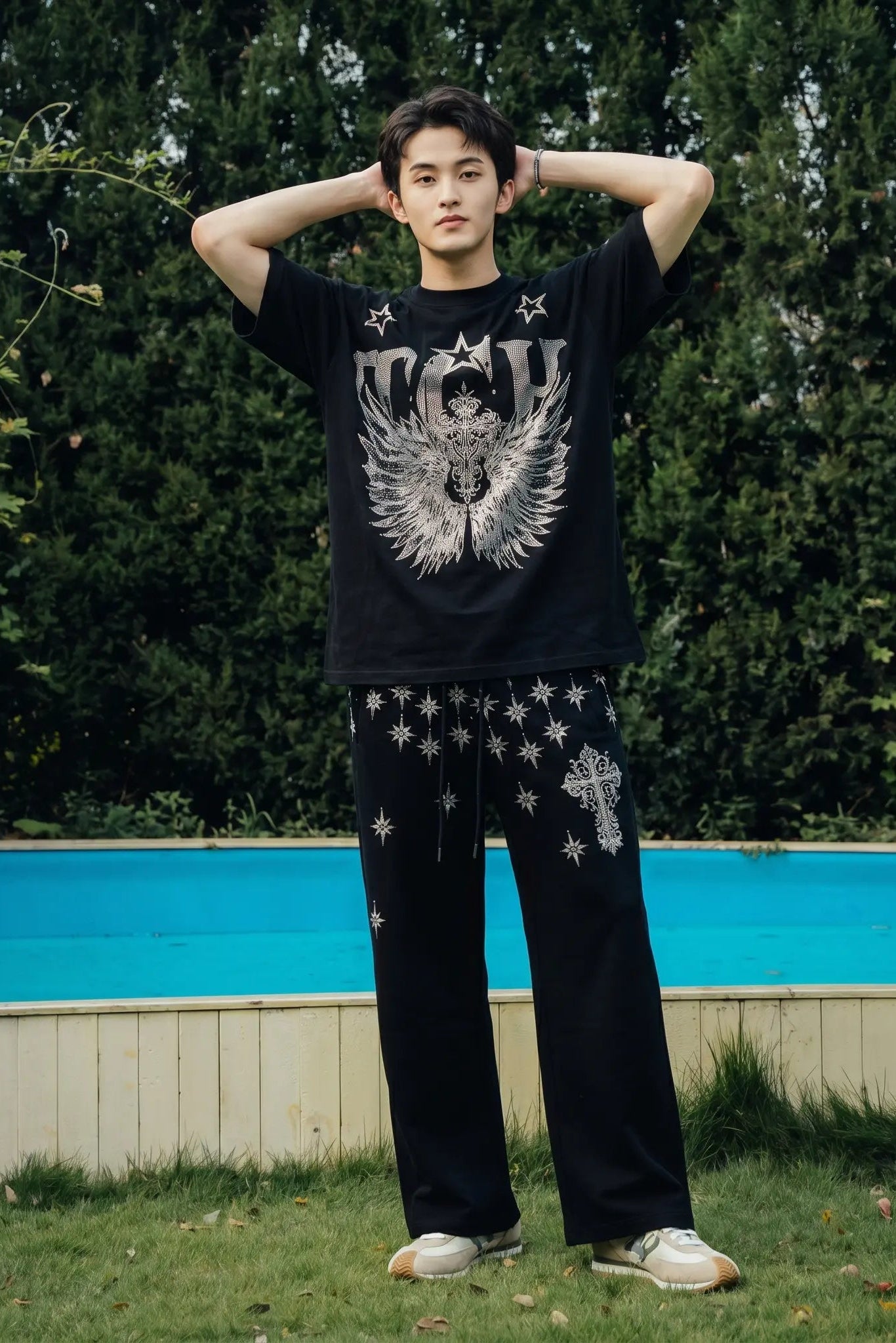 A man stands outdoors in front of a blue pool and green trees, wearing a black graphic T-shirt with a large white angel design and stars, paired with black pants featuring white star prints and white sneakers. TCH Silver Wing Techno Oversized Graphic T-Shirt