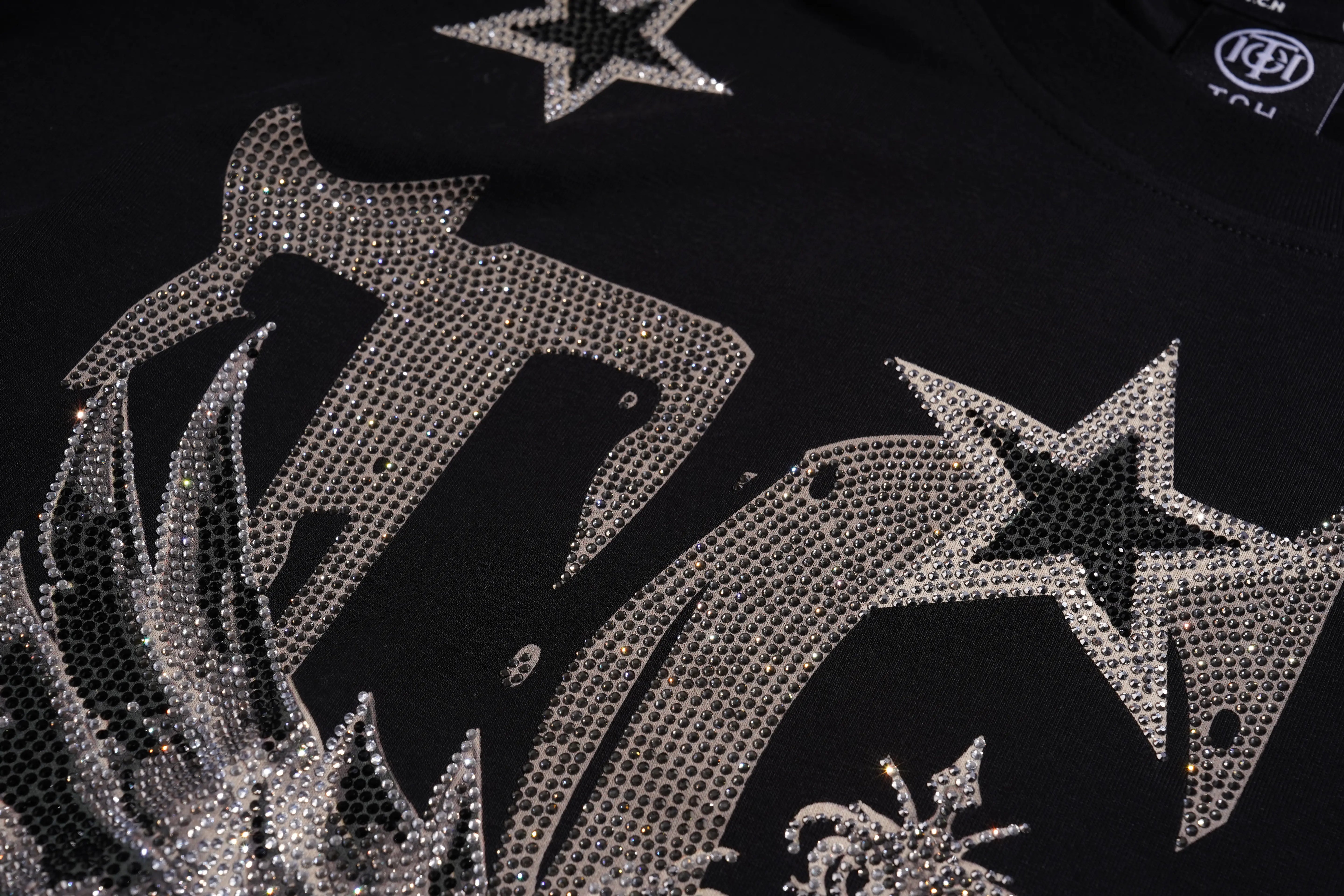 Close-up of a black garment featuring large silver rhinestone star and abstract graphic embellishments, with a small circular logo visible near the collar. TCH Silver Wing Techno Oversized Graphic T-Shirt