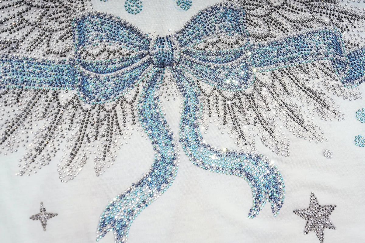 A close-up of a white fabric featuring a large angel wing design embellished with light blue, silver, and gray rhinestones, with small silver star accents. TCH Small Bear Angel Wings Rhinestone Graphic White Long Sleeve T-Shirt