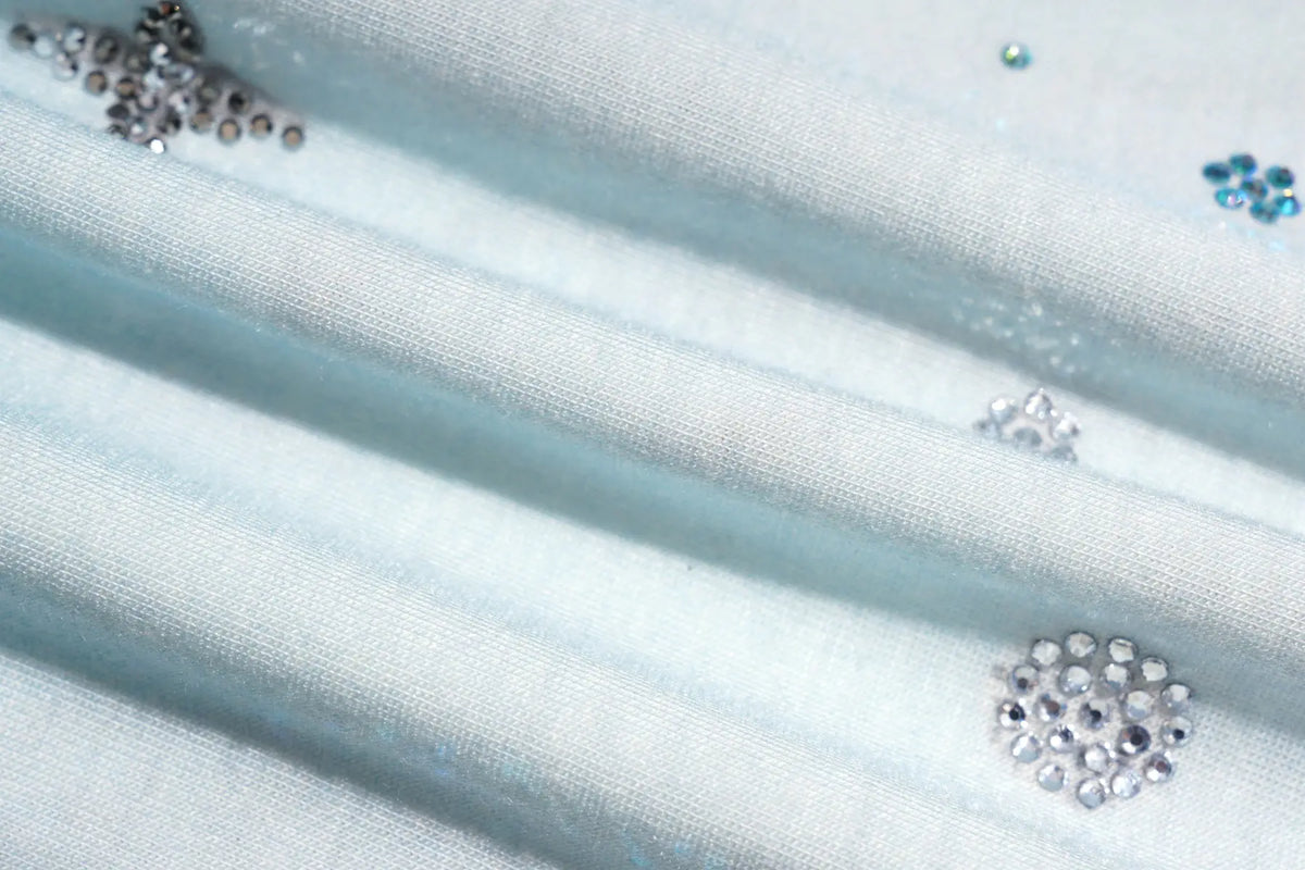 A close-up of light blue fabric with silver and clear rhinestone embellishments arranged in decorative clusters. TCH Small Bear Angel Wings Rhinestone Graphic White Long Sleeve T-Shirt