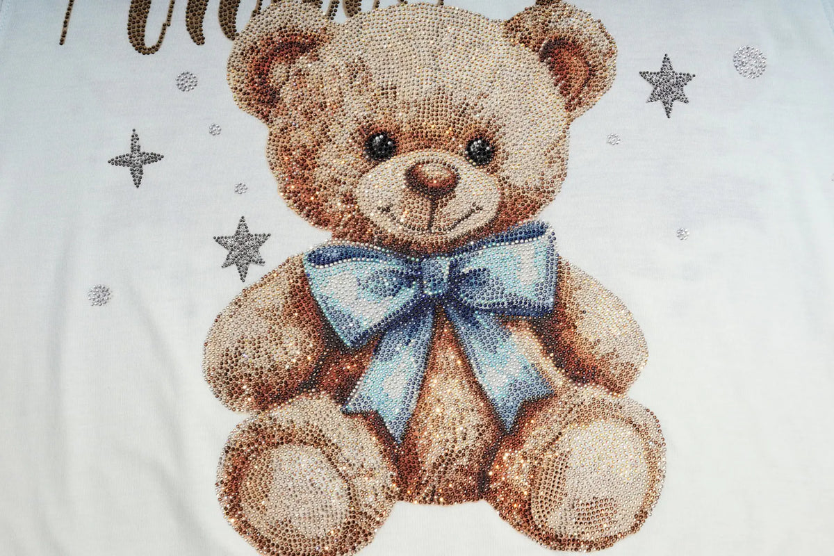 A close-up of a white fabric surface featuring a teddy bear design made from small rhinestones. The bear is light brown with a light blue bow and silver star embellishments. TCH Small Bear Angel Wings Rhinestone Graphic White Long Sleeve T-Shirt