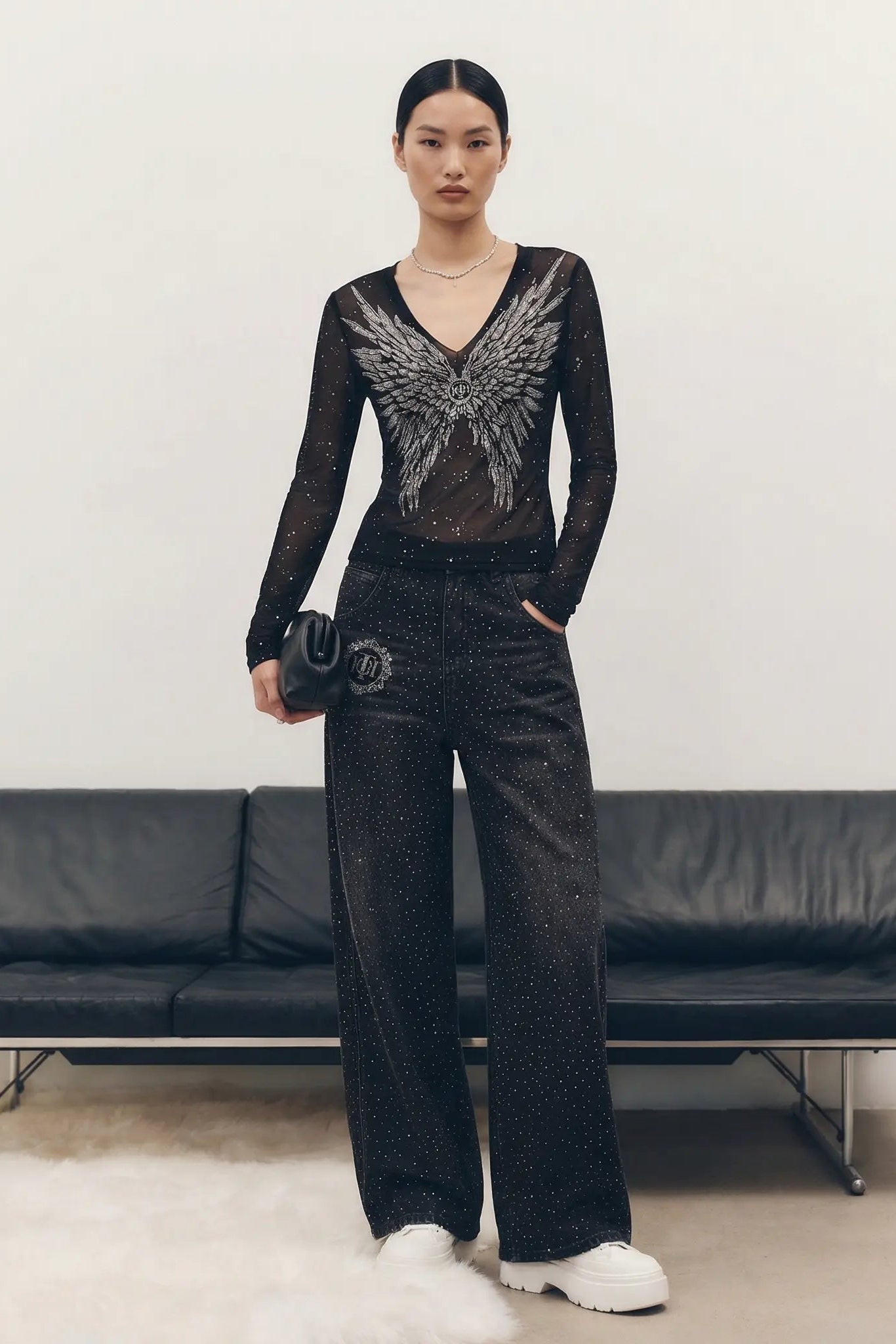 Model wearing a black outfit with a detailed design, standing in front of a black leather couch. TCH Starry Rhinestone Logo Black Denim Jeans