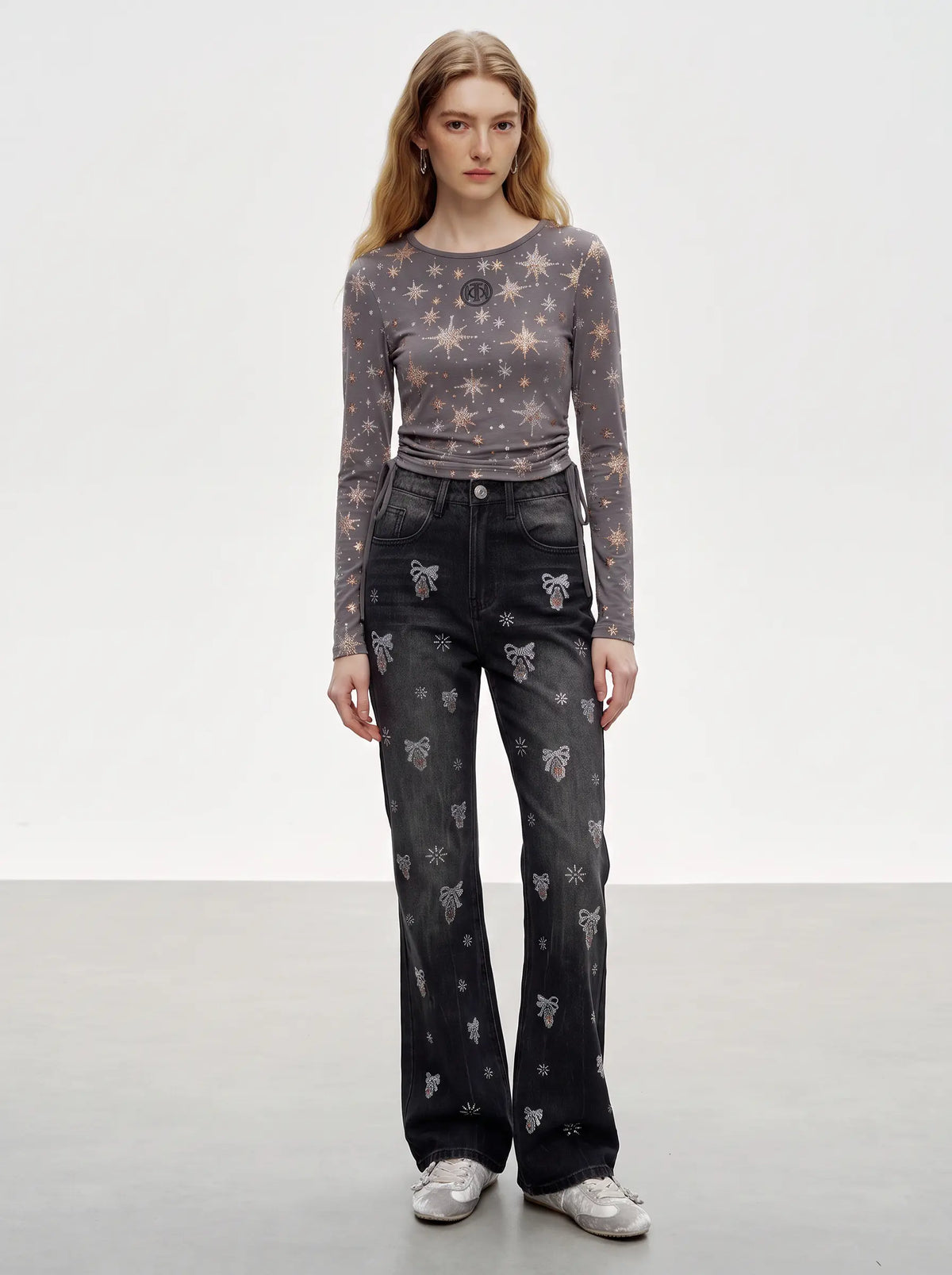 A model stands against a light gray studio background wearing a sheer long-sleeve top with a star pattern, dark gray jeans with butterfly prints, and silver sneakers.