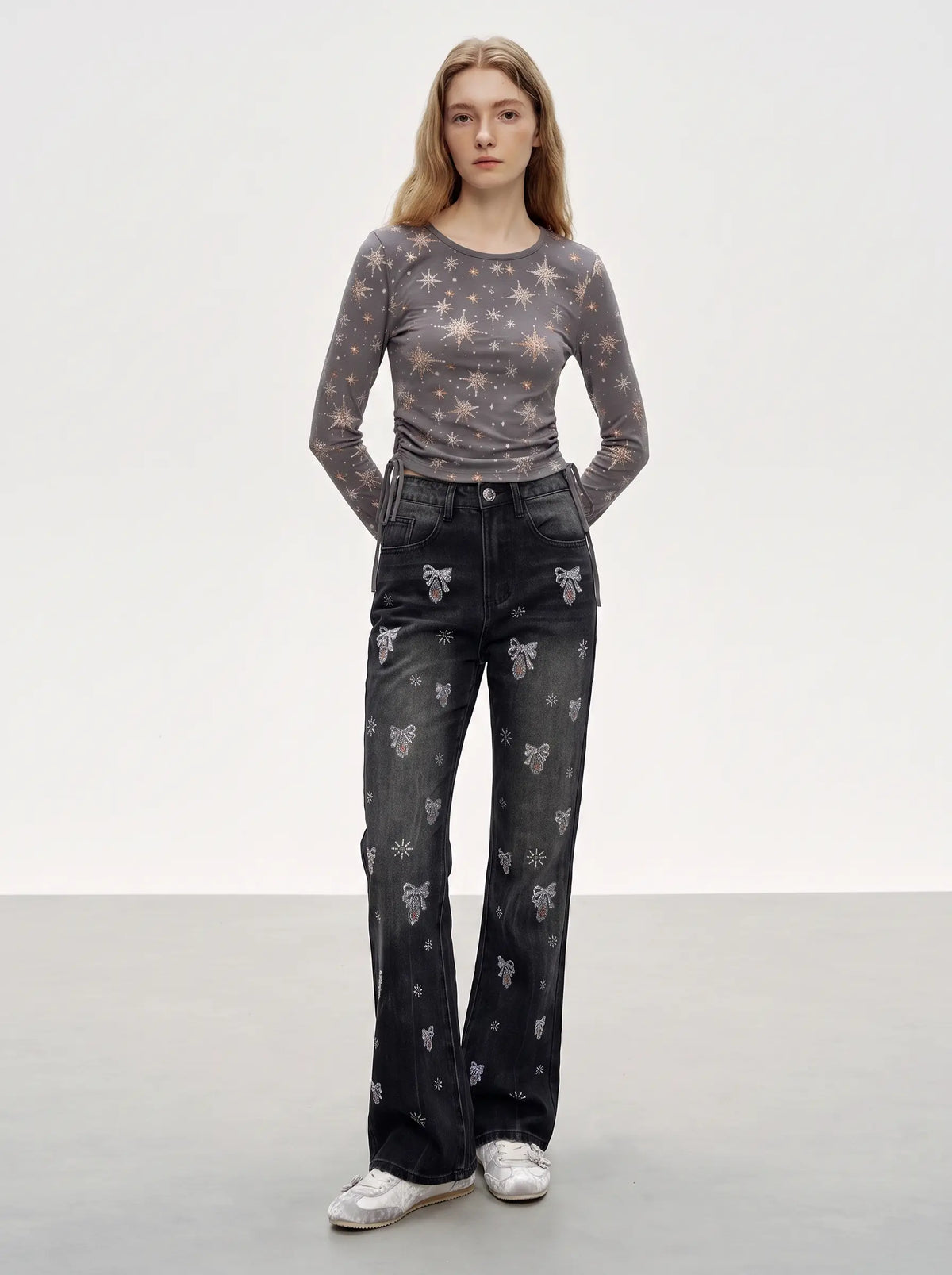 A model stands in a studio wearing a sheer long-sleeve top with a star pattern, paired with dark wide-leg jeans featuring butterfly prints and white sneakers. TCH Starry Rhinestone Print Ruched Long Sleeve Top