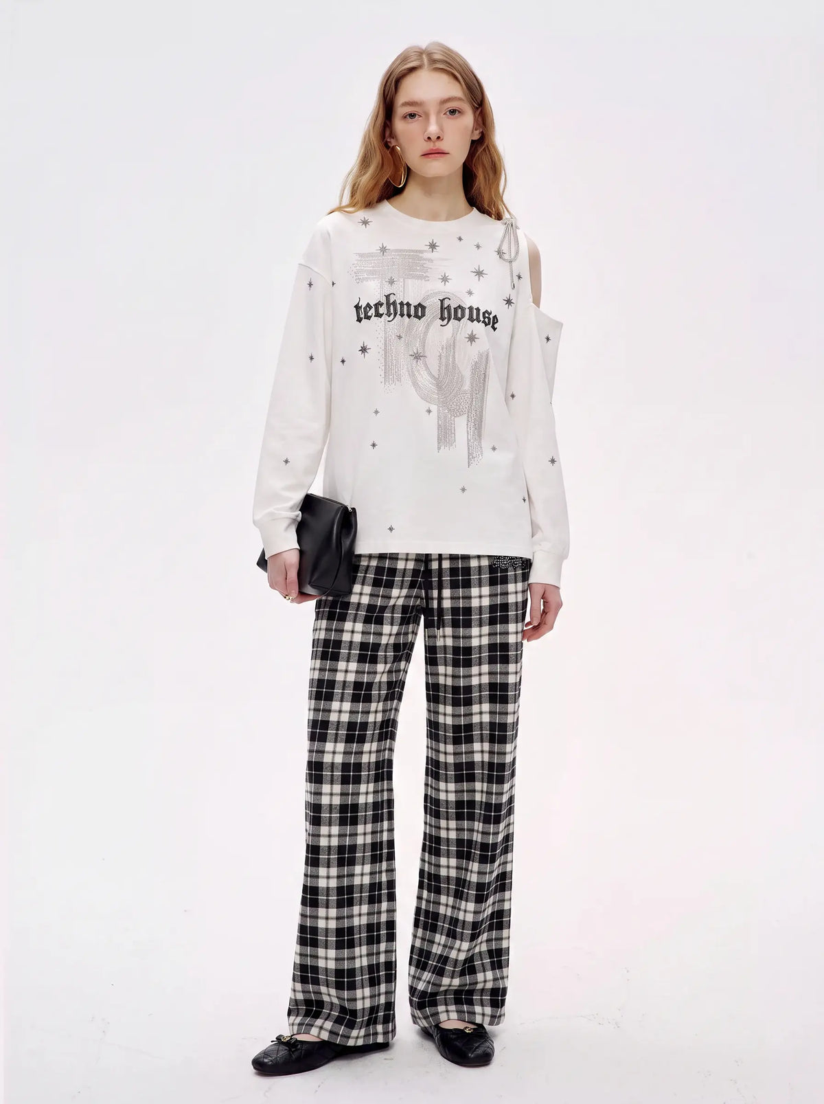 A model stands against a plain white studio background wearing a white long-sleeve sweatshirt with 'techno house' text and a black-and-white plaid pair of wide-leg pants. TCH Starry Western Graphic White Long Sleeve Top