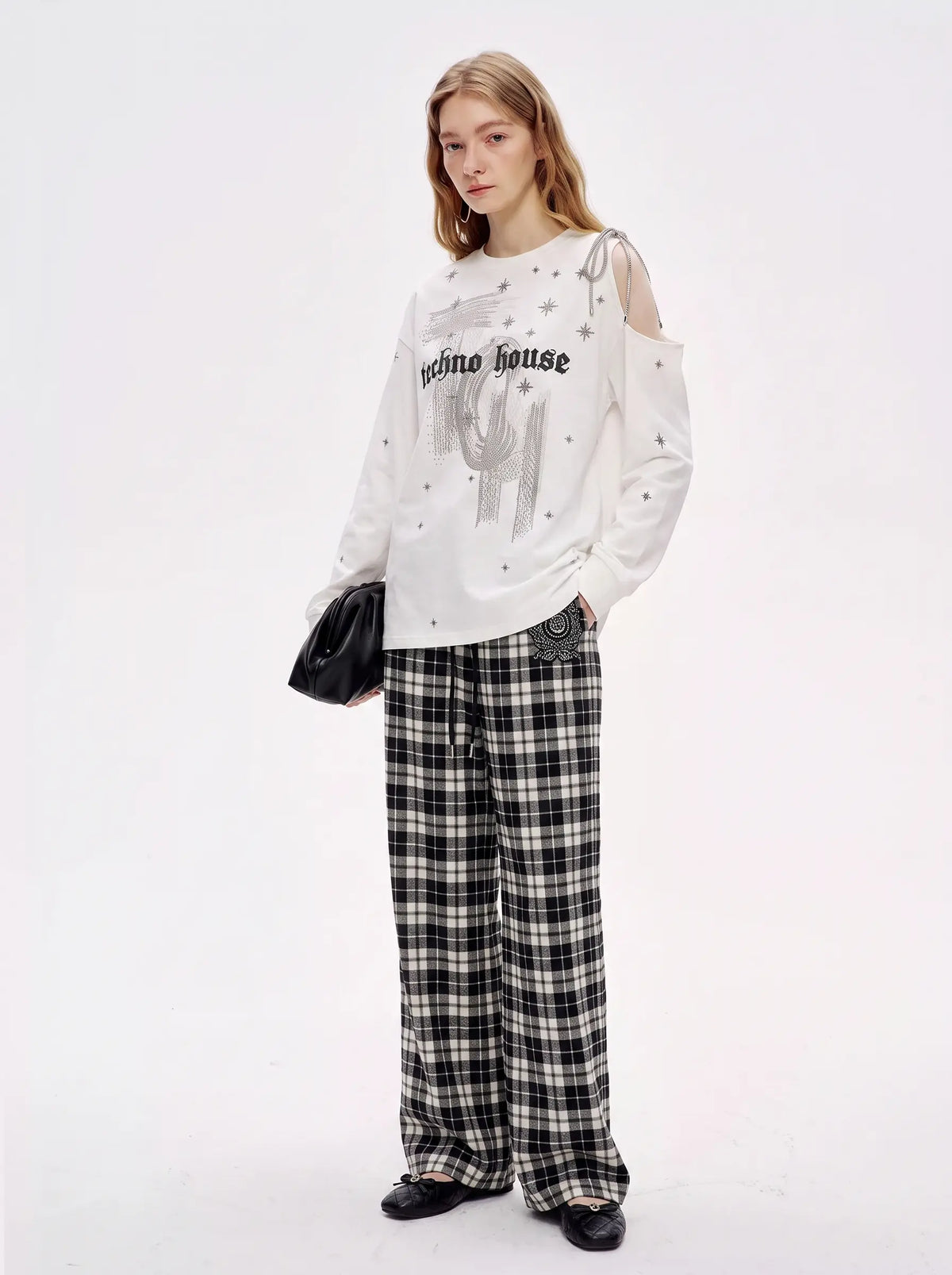 A model stands against a plain white studio background wearing a distressed white long-sleeve graphic top with 'primo house' text and a cactus print, paired with black-and-white plaid wide-leg pants and black shoes. TCH Starry Western Graphic White Long Sleeve Top