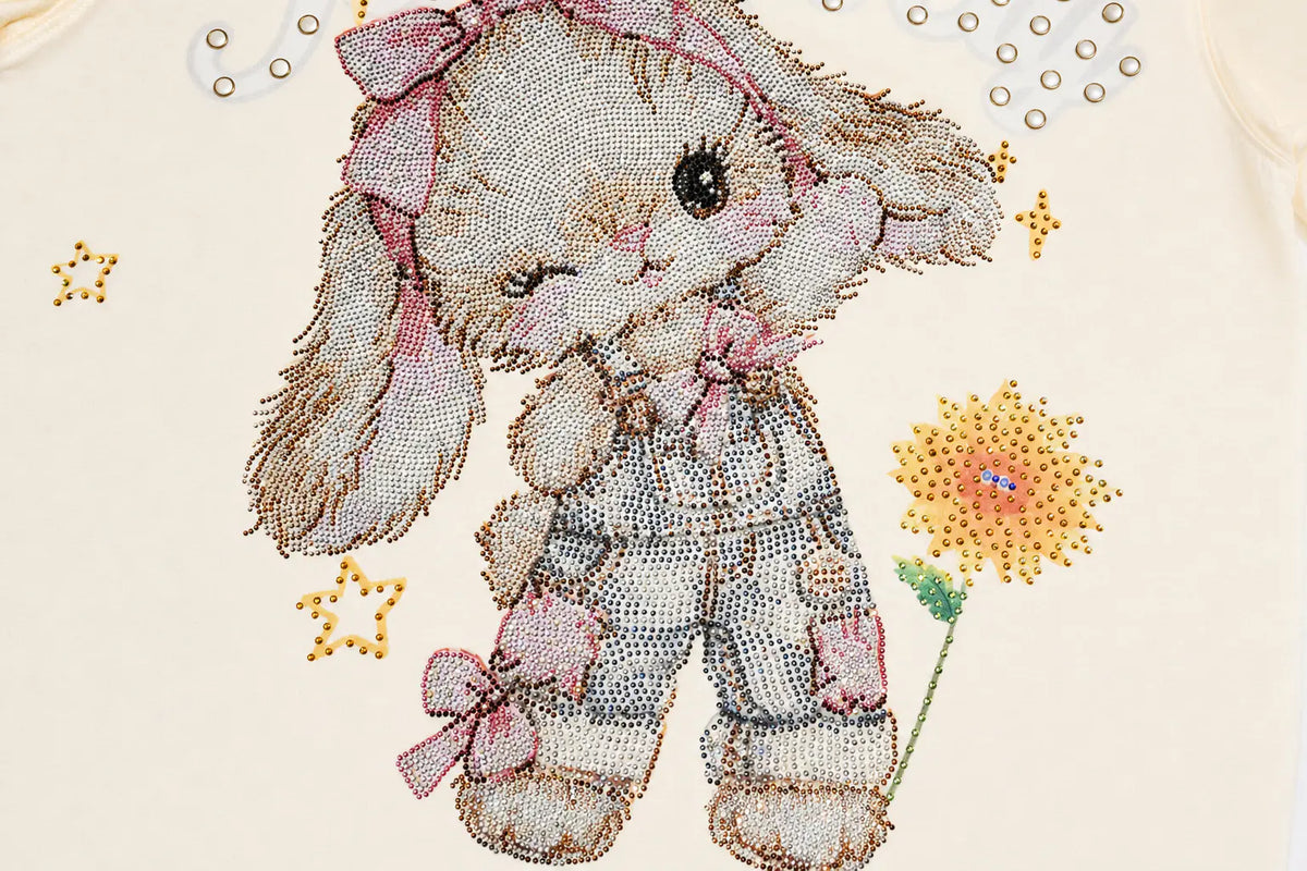 A close-up of a cream-colored beaded craft kit design featuring a cute bunny character wearing pink bows and striped pajamas, holding a yellow flower, with small gold star embellishments. TCH Sweet Bunny Rhinestone Graphic Long Sleeve Top