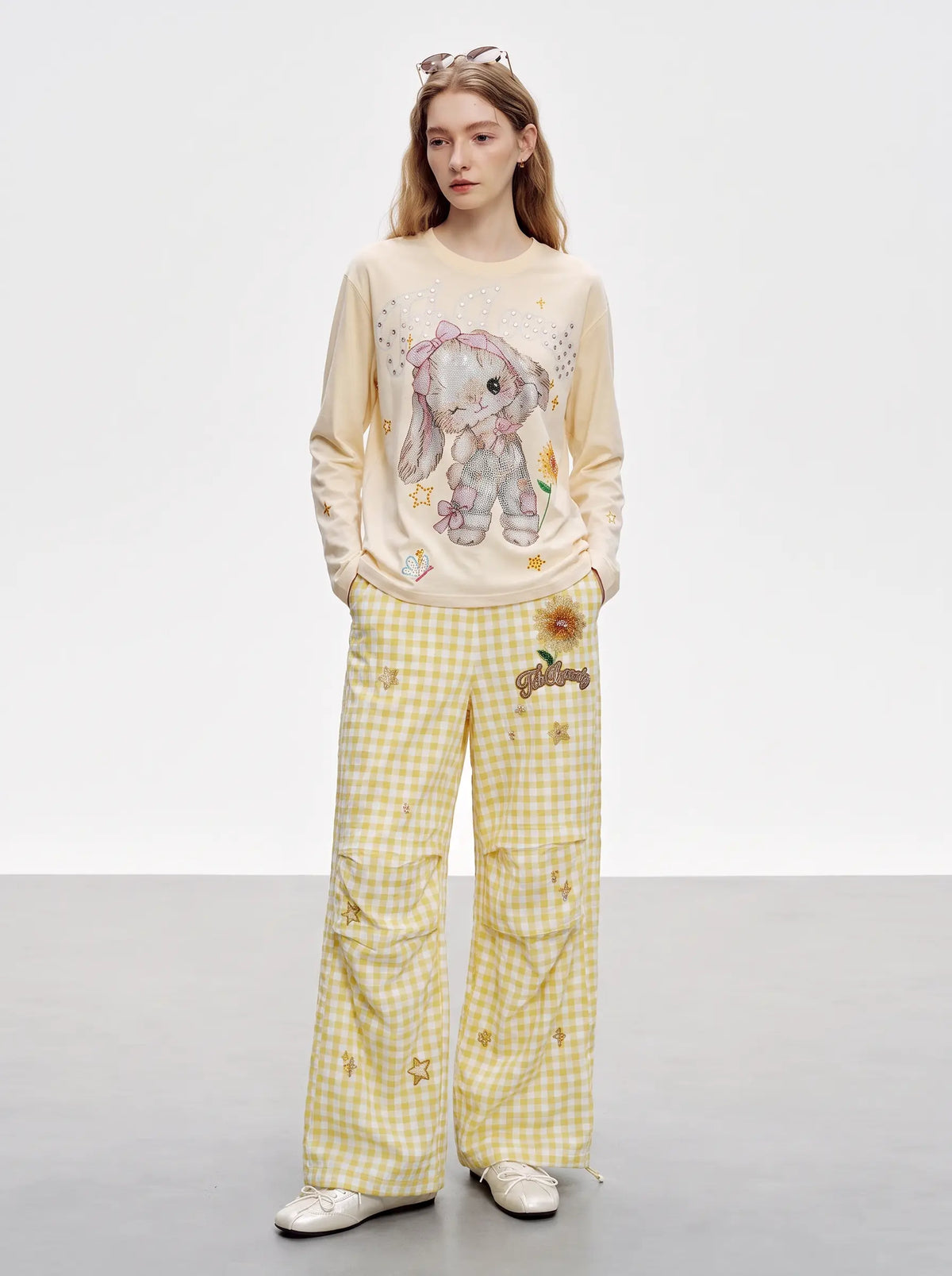 A model stands against a light gray studio background wearing a cream long-sleeve sweatshirt with a bunny graphic and yellow-and-white gingham pants with small embroidered details. TCH Sweet Bunny Rhinestone Graphic Long Sleeve Top