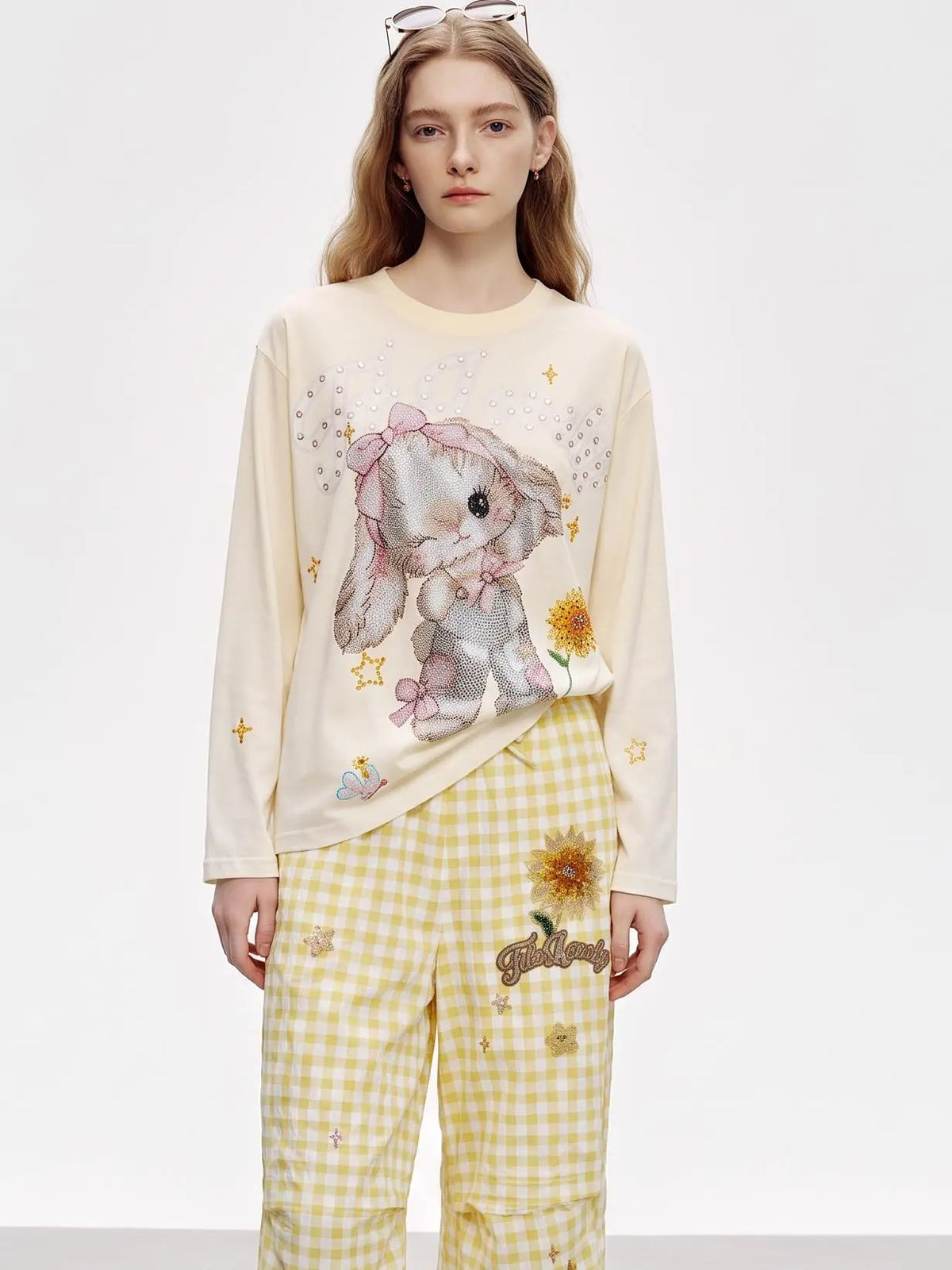 A model wearing a cream-colored long-sleeve t-shirt with a bunny graphic and yellow-and-white gingham pants with a sunflower patch. TCH Sweet Bunny Rhinestone Graphic Long Sleeve Top