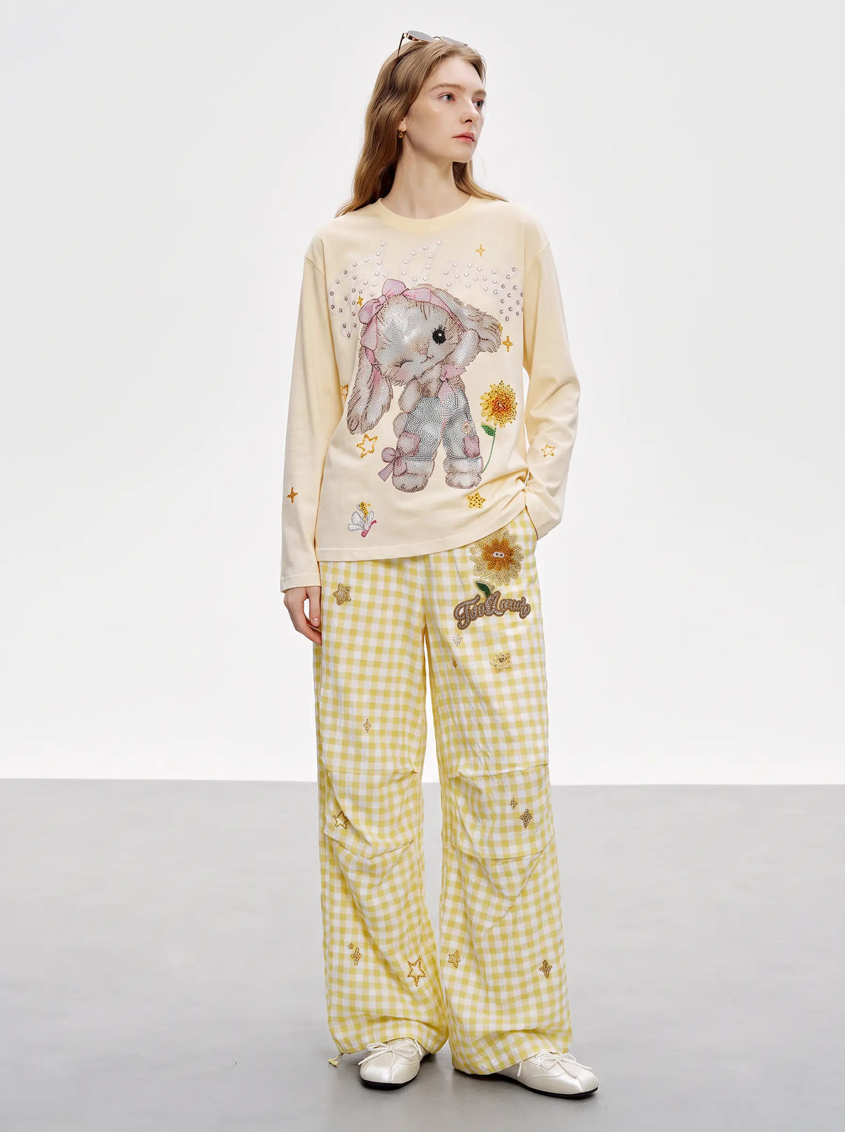 A model wearing a two-piece pajama set consisting of a cream long-sleeve top with a bunny graphic and yellow-and-white gingham pants. TCH Sweet Bunny Rhinestone Graphic Long Sleeve Top