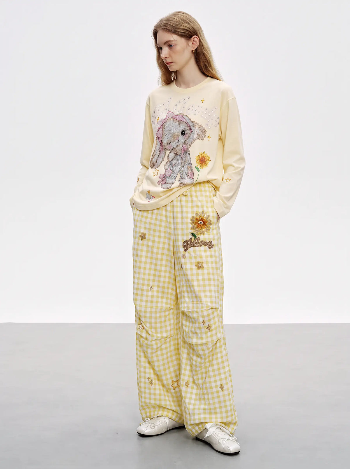 A model stands in a studio wearing a cream-colored long-sleeve top with a bunny graphic and yellow-and-white gingham wide-leg pants with floral embroidery. TCH Sweet Bunny Rhinestone Graphic Long Sleeve Top