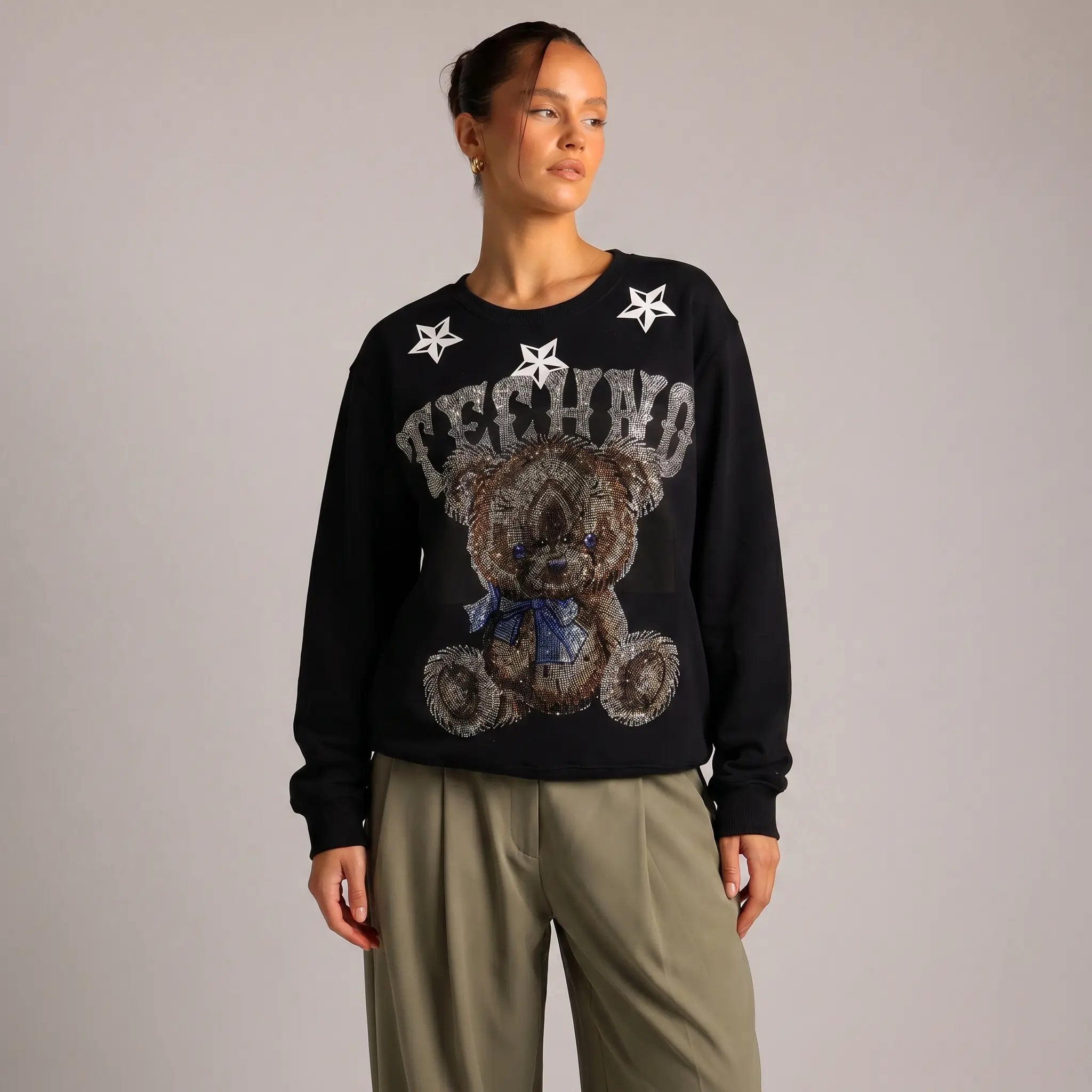 Person wearing a black sweater with a teddy bear design and 'Techno' text on a plain background, TCH TECHNO Rhinestone Teddy Sweatshirt