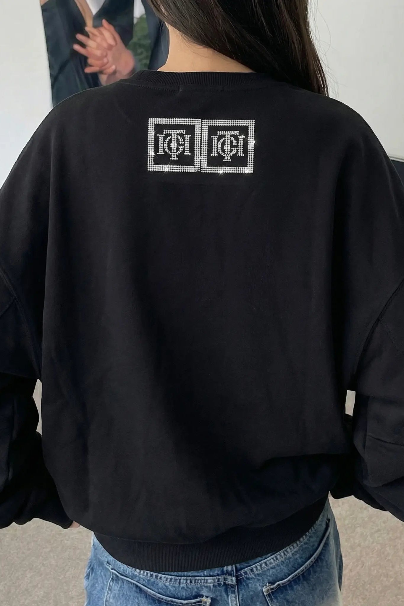 Person wearing a black sweatshirt with a logo on the back, standing indoors. TCH TECHNO Rhinestone Teddy Sweatshirt