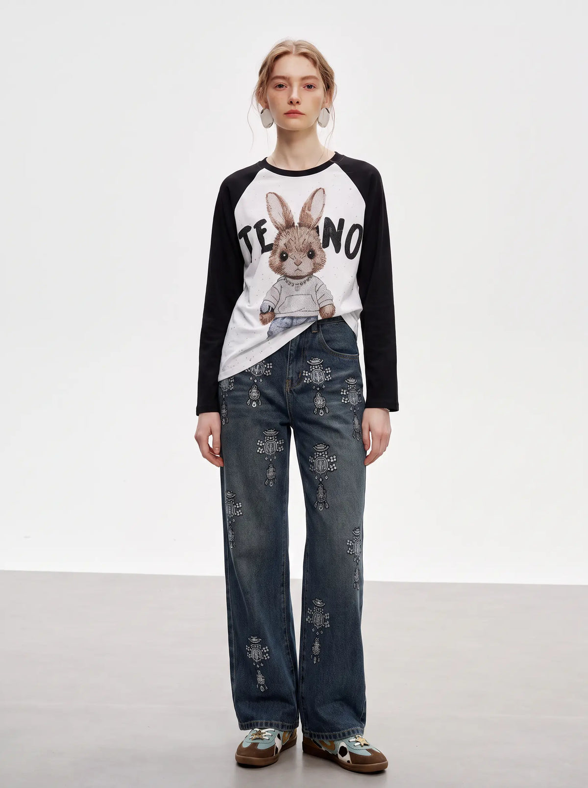 A model stands against a light gray studio background wearing a raglan-style long-sleeve shirt with a rabbit graphic and text on the front, paired with distressed blue jeans and brown shoes. TCH Techno Bunny Rhinestone Raglan Long Sleeve T-Shirt