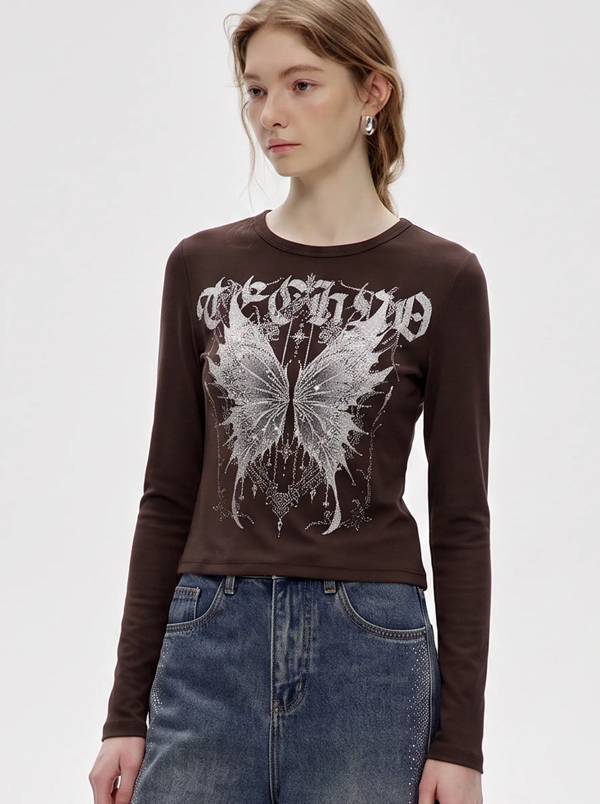 A model wearing a light blue denim jacket with various embroidered patches, including a teddy bear on the left chest and cherries on the sleeve, styled over a white t-shirt and dark pants. TCH Techno Butterfly Starburst Graphic Long Sleeve Top