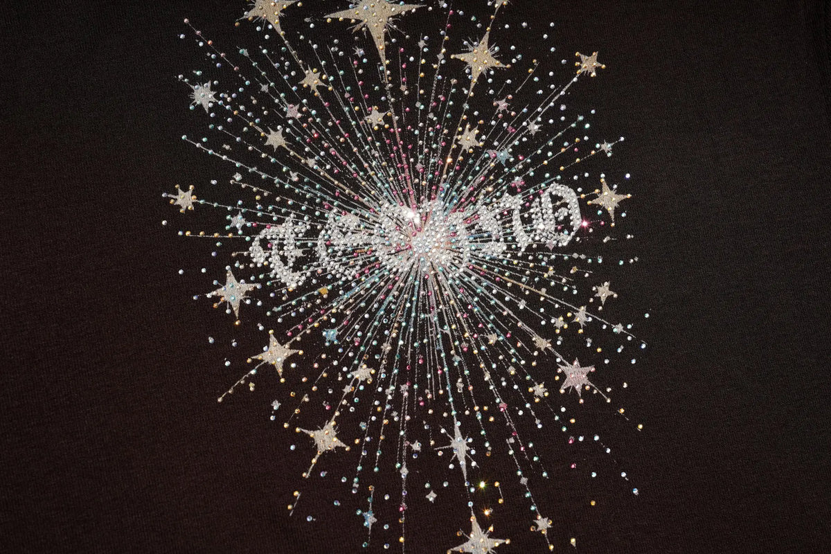 A close-up of a black fabric surface decorated with a glittering starburst design made of rhinestones and iridescent beads, with the word 'celebrate' spelled out in the center. TCH Techno Butterfly Starburst Graphic Long Sleeve Top