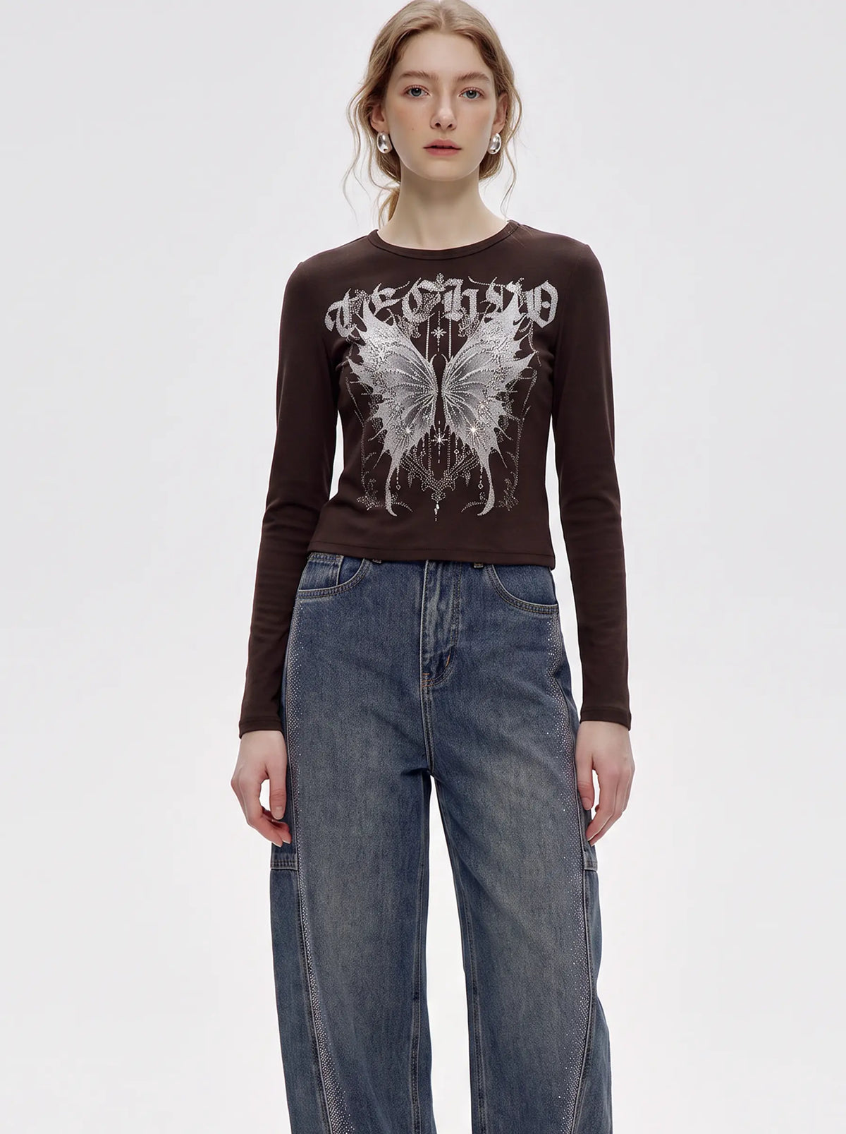 A model wearing a dark brown long-sleeve crop top with a white gothic-style graphic and butterfly print, paired with high-waisted blue jeans, against a plain light gray background. TCH Techno Butterfly Starburst Graphic Long Sleeve Top