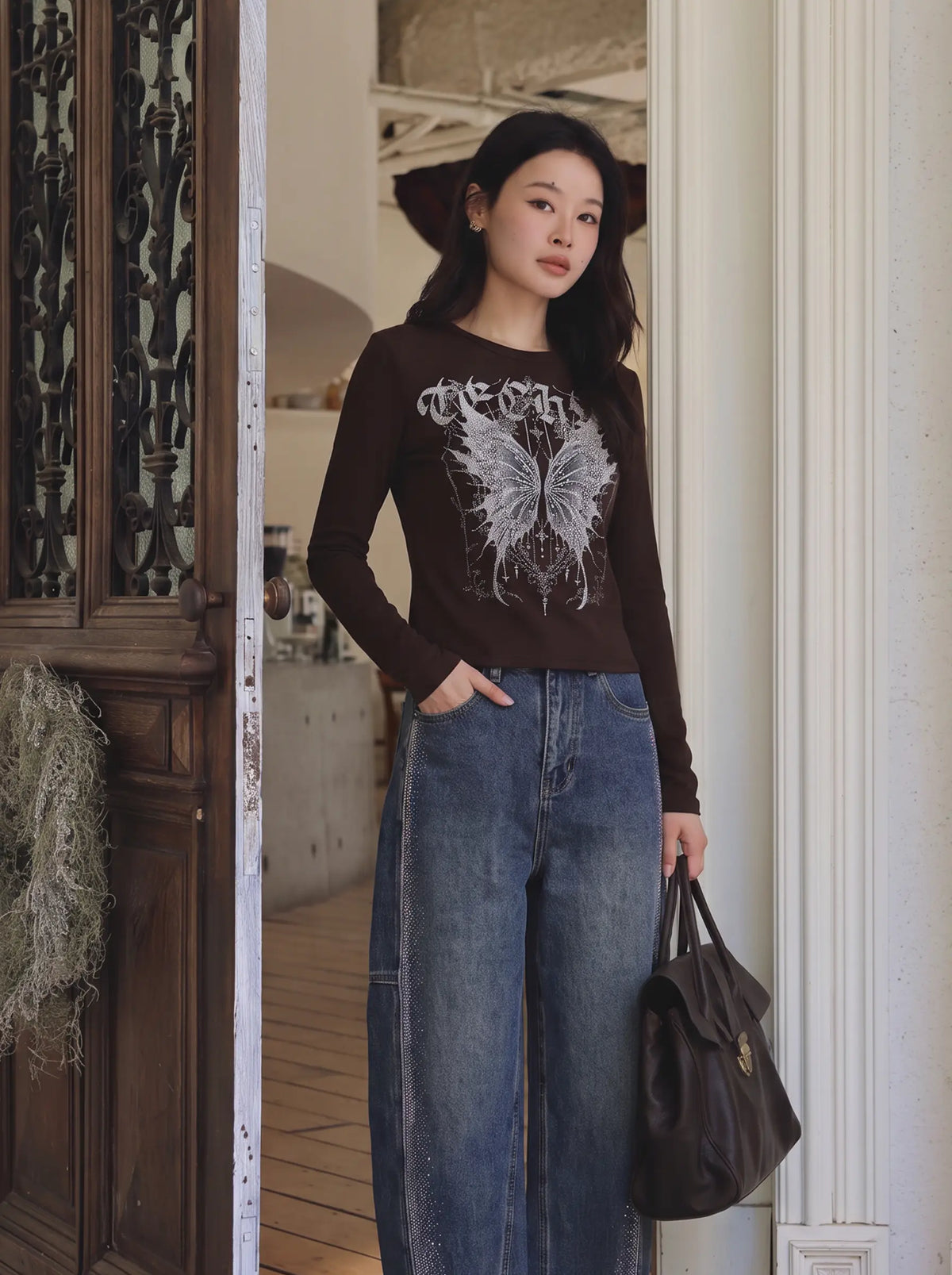 A woman stands in a doorway wearing a dark brown long-sleeve graphic top with a white butterfly print and blue wide-leg jeans, holding a dark brown leather handbag. TCH Techno Butterfly Starburst Graphic Long Sleeve Top
