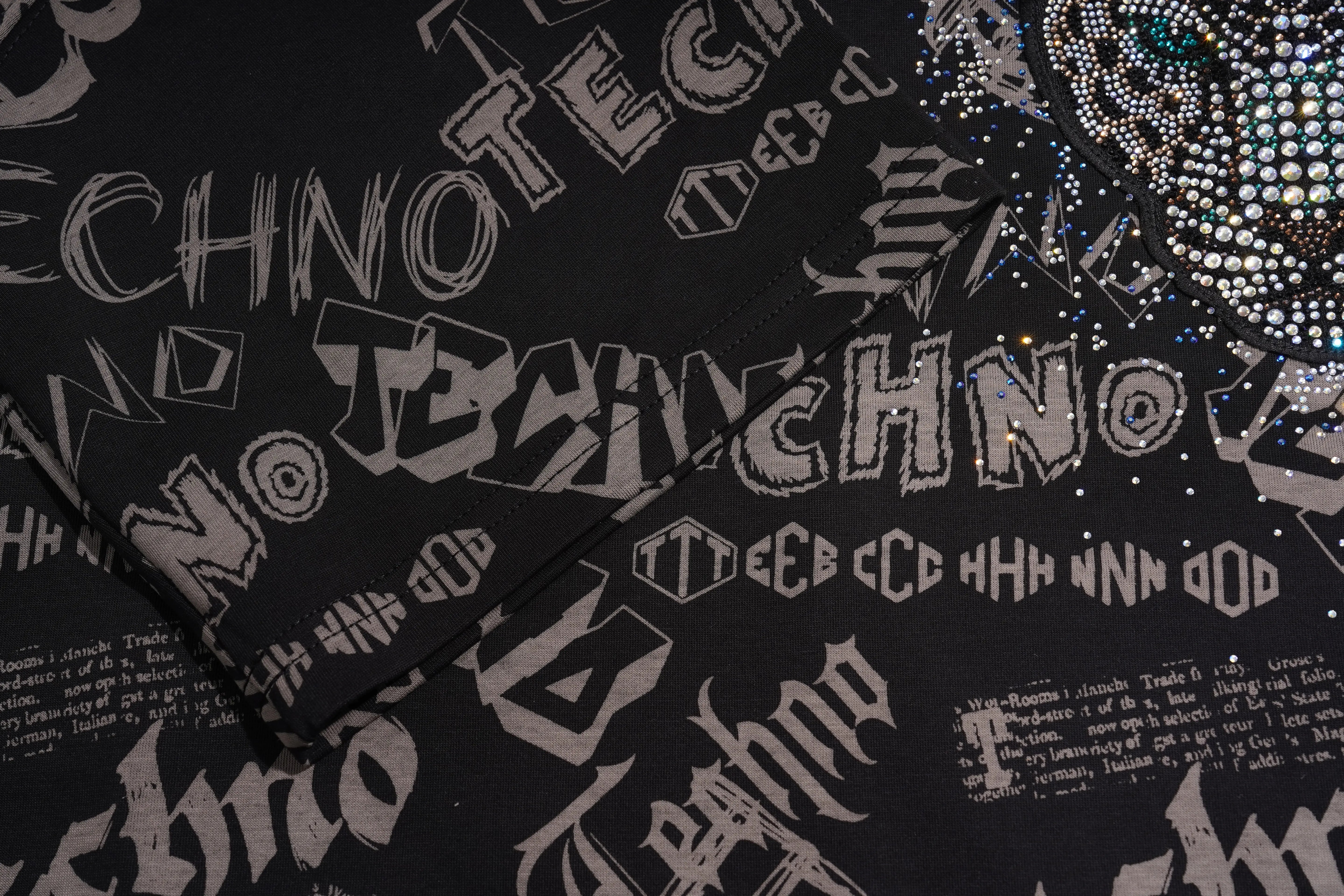 Black fabric with a repeating gothic-style 'TECHNO' graphic print in light gray, overlaid with silver rhinestone embellishments and sequins. TCH Techno Graffiti Rhinestone Skull Oversized T-Shirt