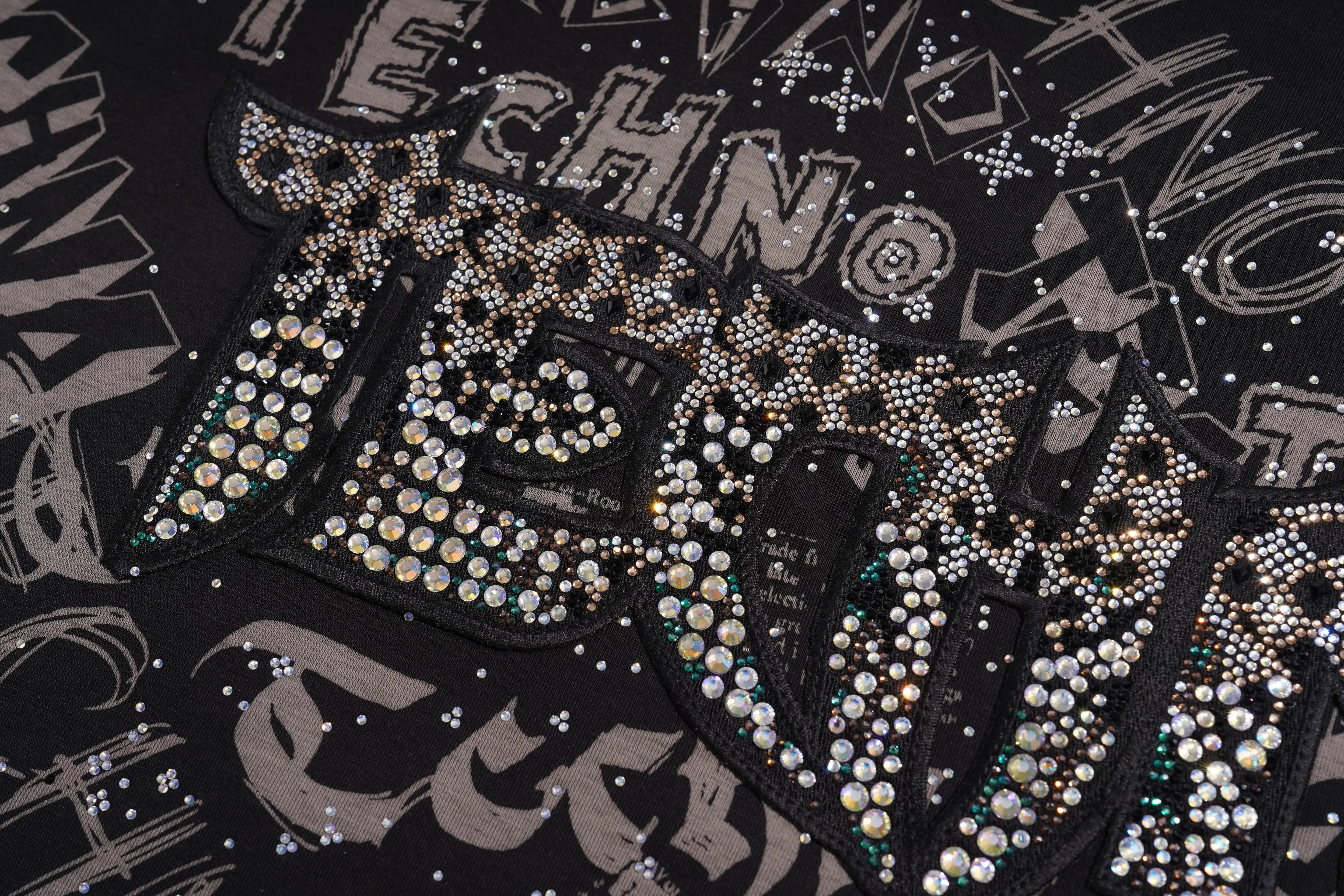 Close-up of a black graphic t-shirt featuring a large, heavily embellished patch with rhinestones spelling 'TECHNO' and abstract lettering on a black background. TCH Techno Graffiti Rhinestone Skull Oversized T-Shirt