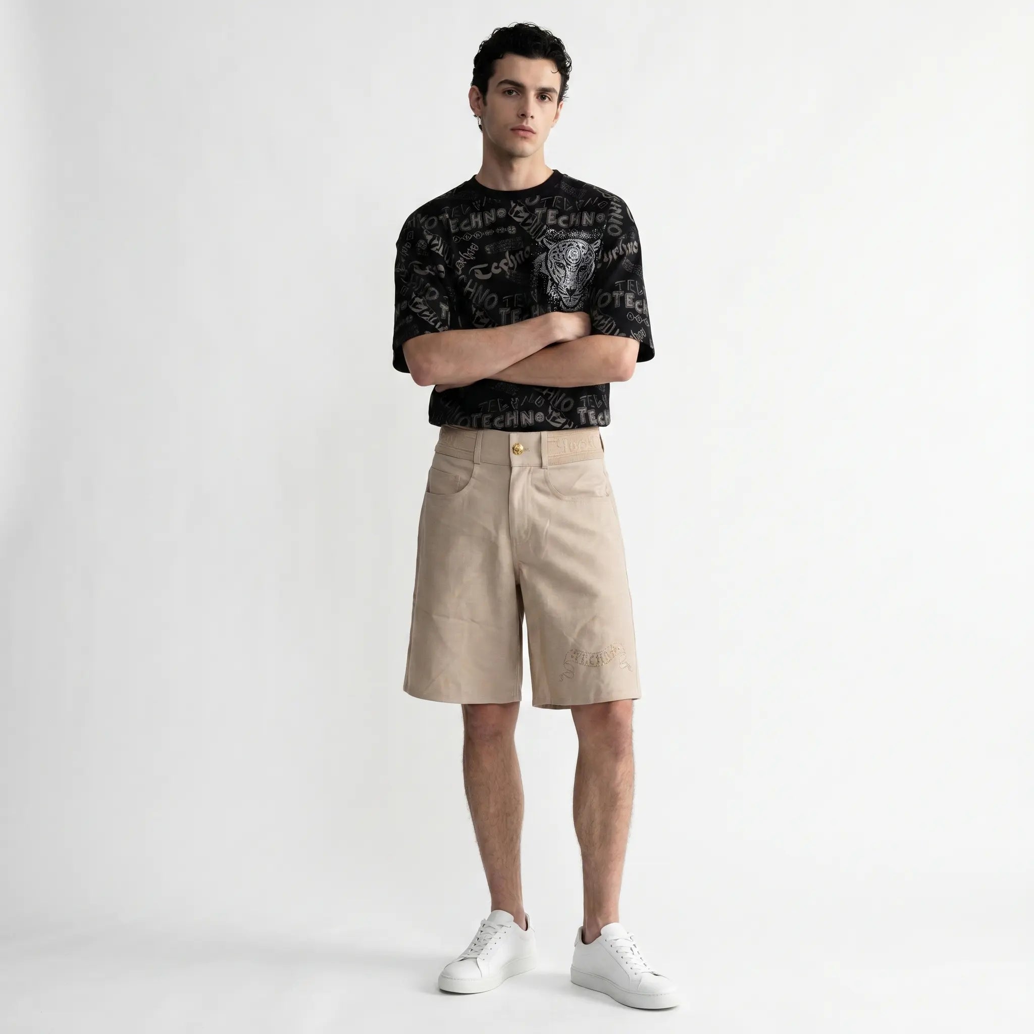 A male model stands against a light gray studio background wearing a black short-sleeve graphic T-shirt, beige shorts, and white sneakers. TCH Techno Graffiti Rhinestone Skull Oversized T-Shirt