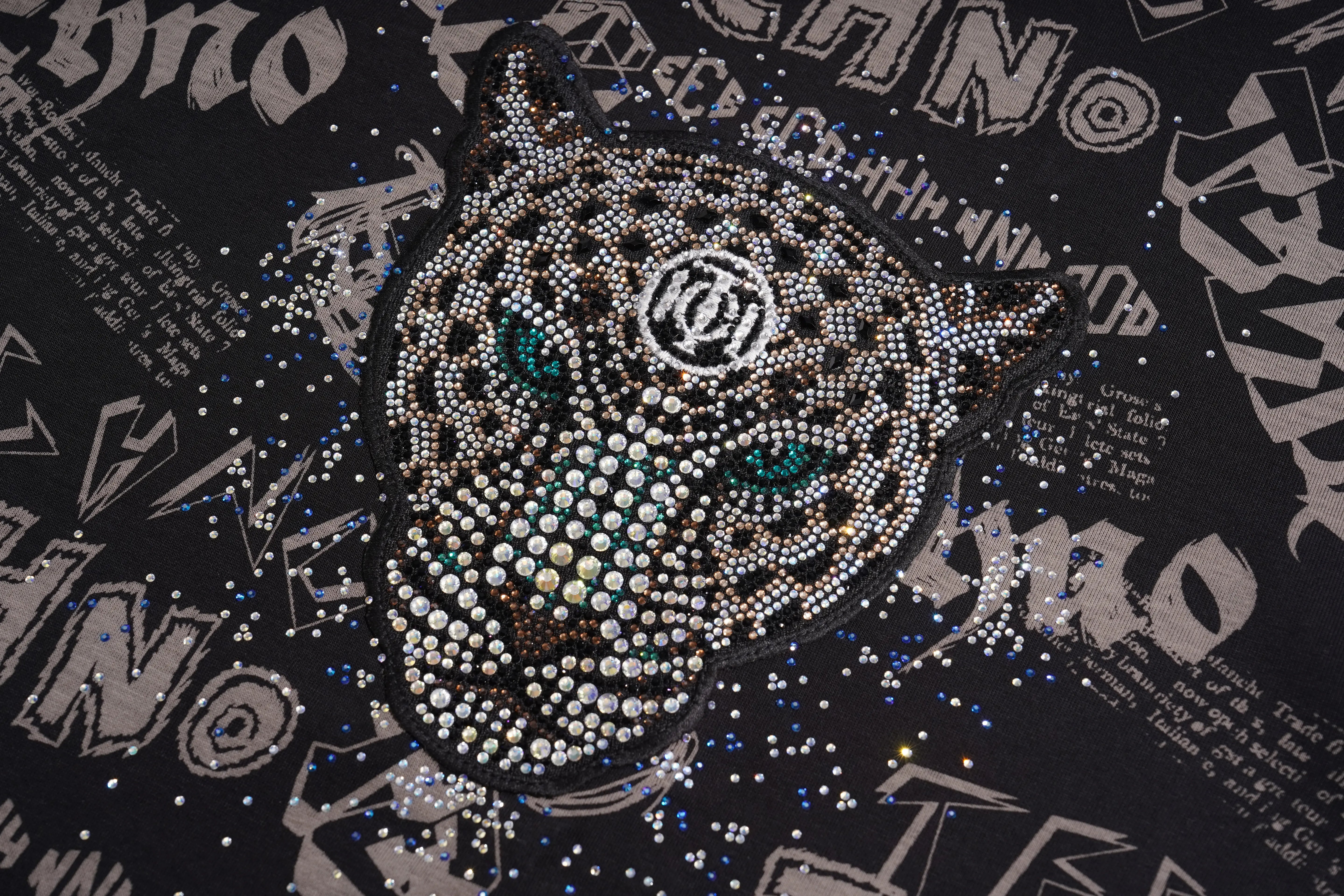 A close-up of a black graphic garment featuring a large rhinestone-embellished panther face with turquoise eyes and a circular 'KF' emblem in the center. The background has a black-and-white graffiti-style print with scattered rhinestone speckles. TCH Techno Graffiti Rhinestone Skull Oversized T-Shirt