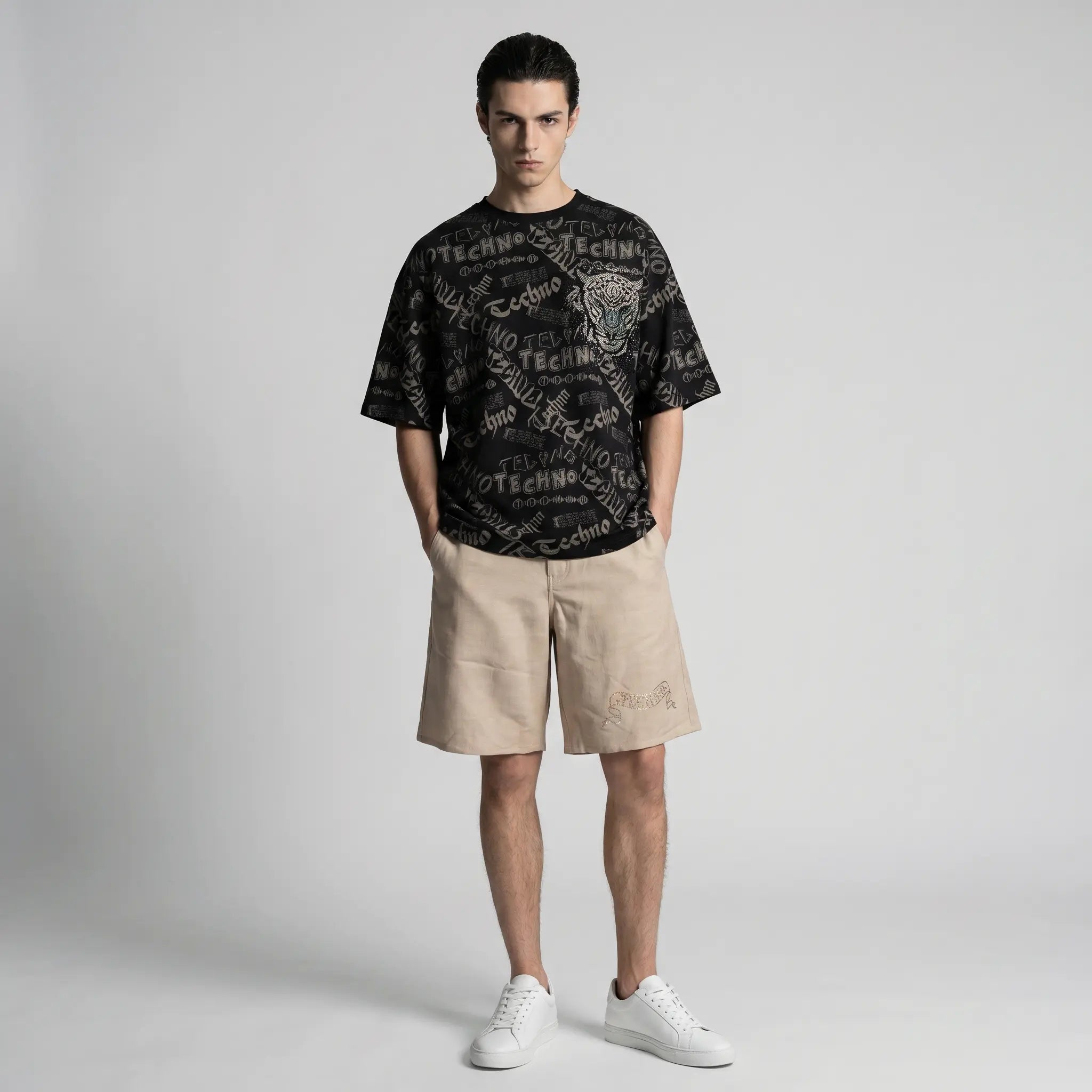 A male model stands against a light gray studio background wearing a black graphic T-shirt with beige shorts and white sneakers. TCH Techno Graffiti Rhinestone Skull Oversized T-Shirt