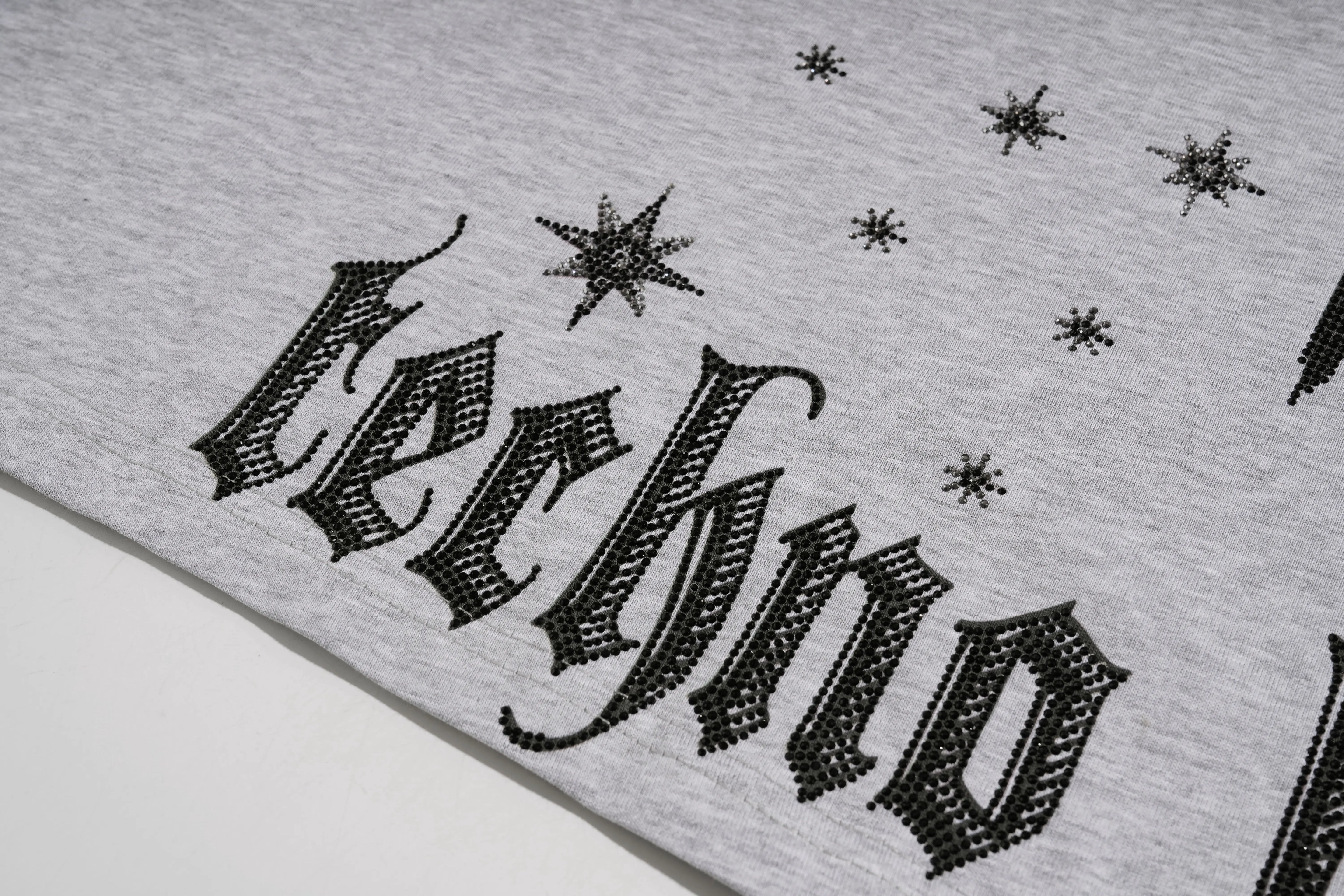 Close-up of a light gray heather fabric printed with the word 'techno' in a black gothic font, decorated with small black starburst motifs. TCH Techno House Abstract Graphic Oversized T-Shirt