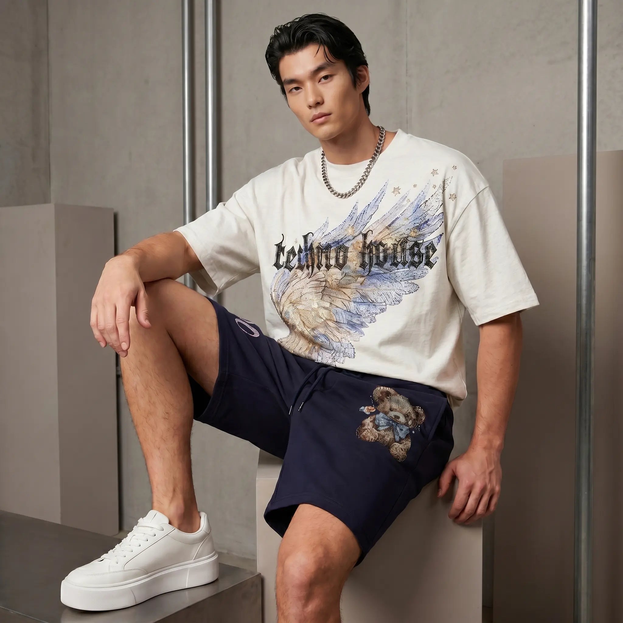 A man models a white graphic t-shirt with 'Leather House' text and angel wings, paired with navy blue shorts featuring a teddy bear graphic and white platform sneakers, seated against a modern concrete wall. TCH Techno House Wing Graphic Oversized T-Shirt