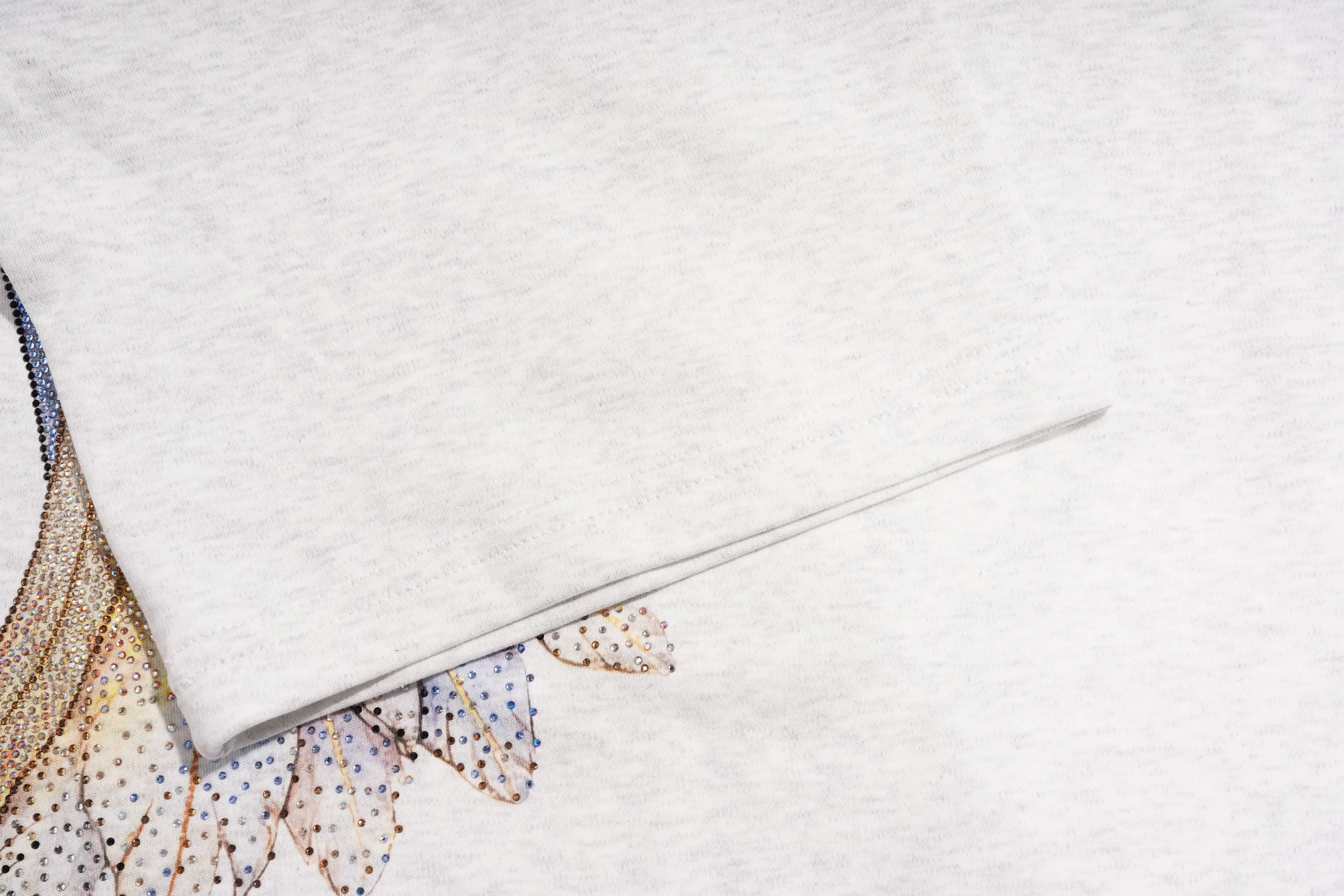 A close-up of a light heather gray fabric, likely a t-shirt, folded over to reveal a detailed graphic print in the bottom-left corner. The print features delicate, patterned wings in shades of blue, gold, and brown with tiny polka dots. TCH Techno House Wing Graphic Oversized T-Shirt