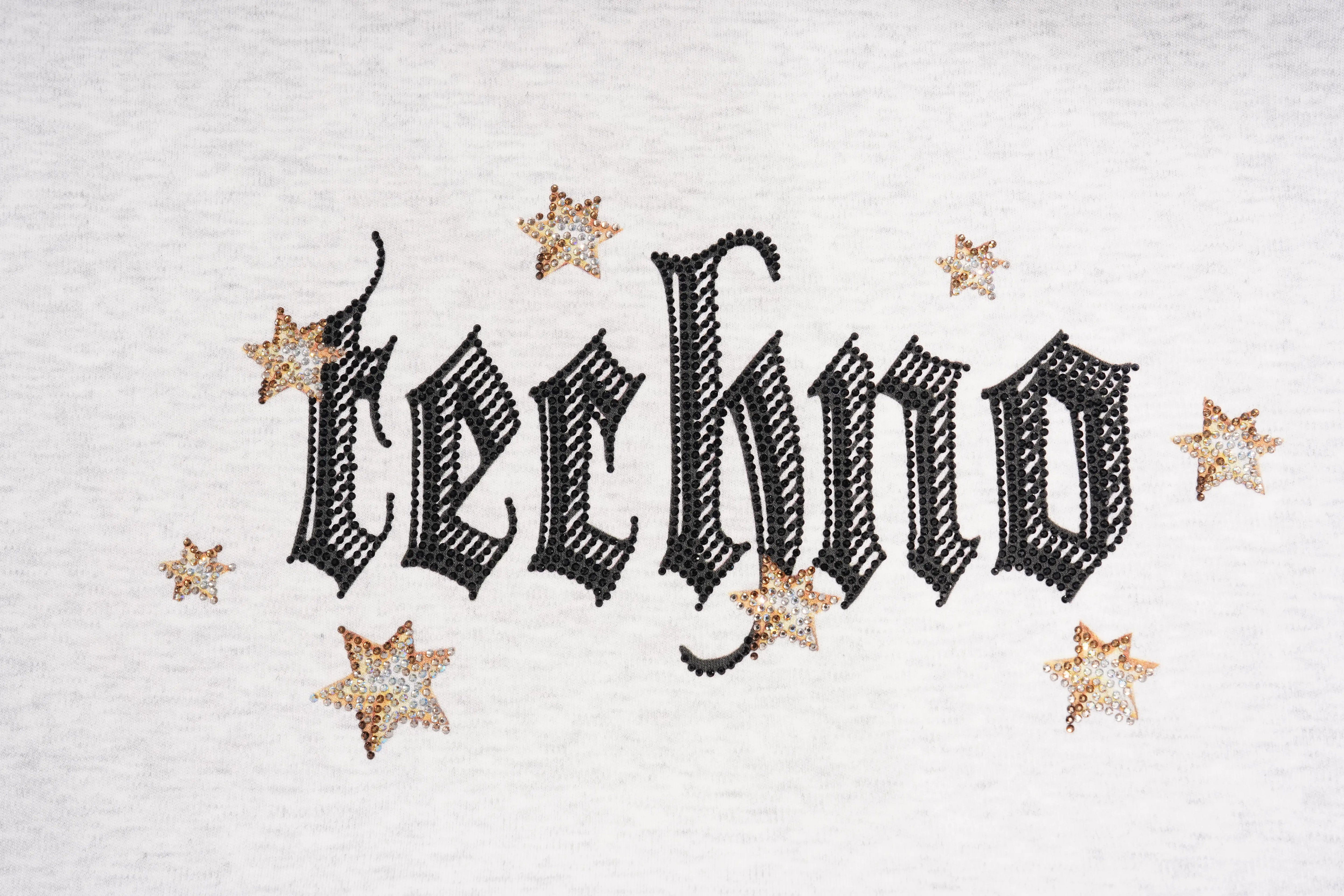 A close-up of a white textured fabric featuring the word 'techno' in a black gothic-style font, decorated with small gold glittery stars. TCH Techno House Wing Graphic Oversized T-Shirt