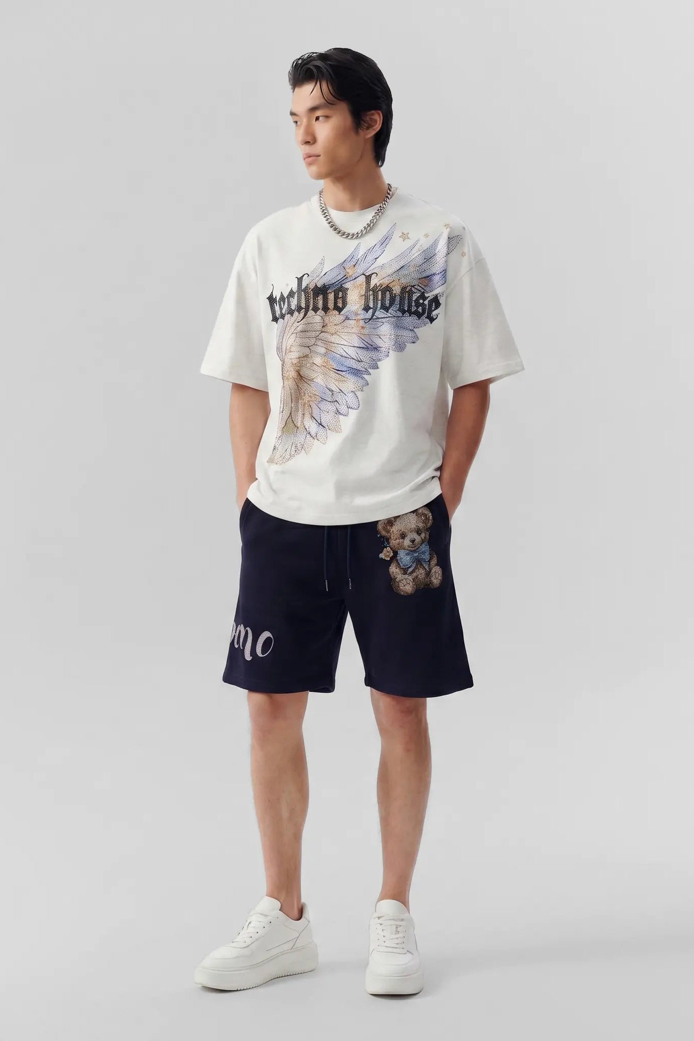 A male model stands against a light gray studio background wearing a white graphic T-shirt with a large angel-wing print and black shorts with a small lion graphic on the thigh. TCH Techno House Wing Graphic Oversized T-Shirt
