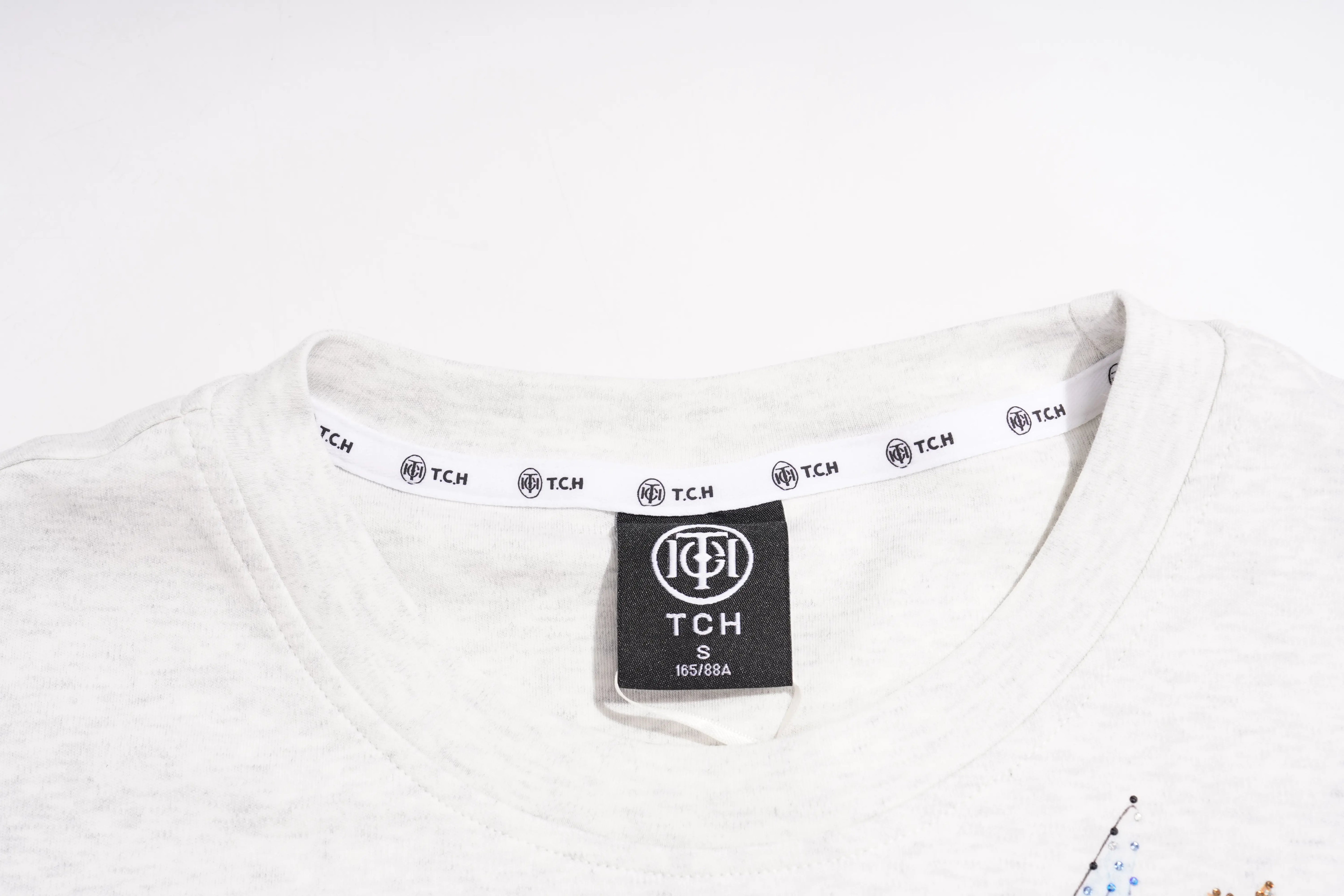 Close-up of a light heather gray crewneck t-shirt showing the inner collar with a white printed TCH brand tag and a black sewn-in neck label. TCH Techno House Wing Graphic Oversized T-Shirt