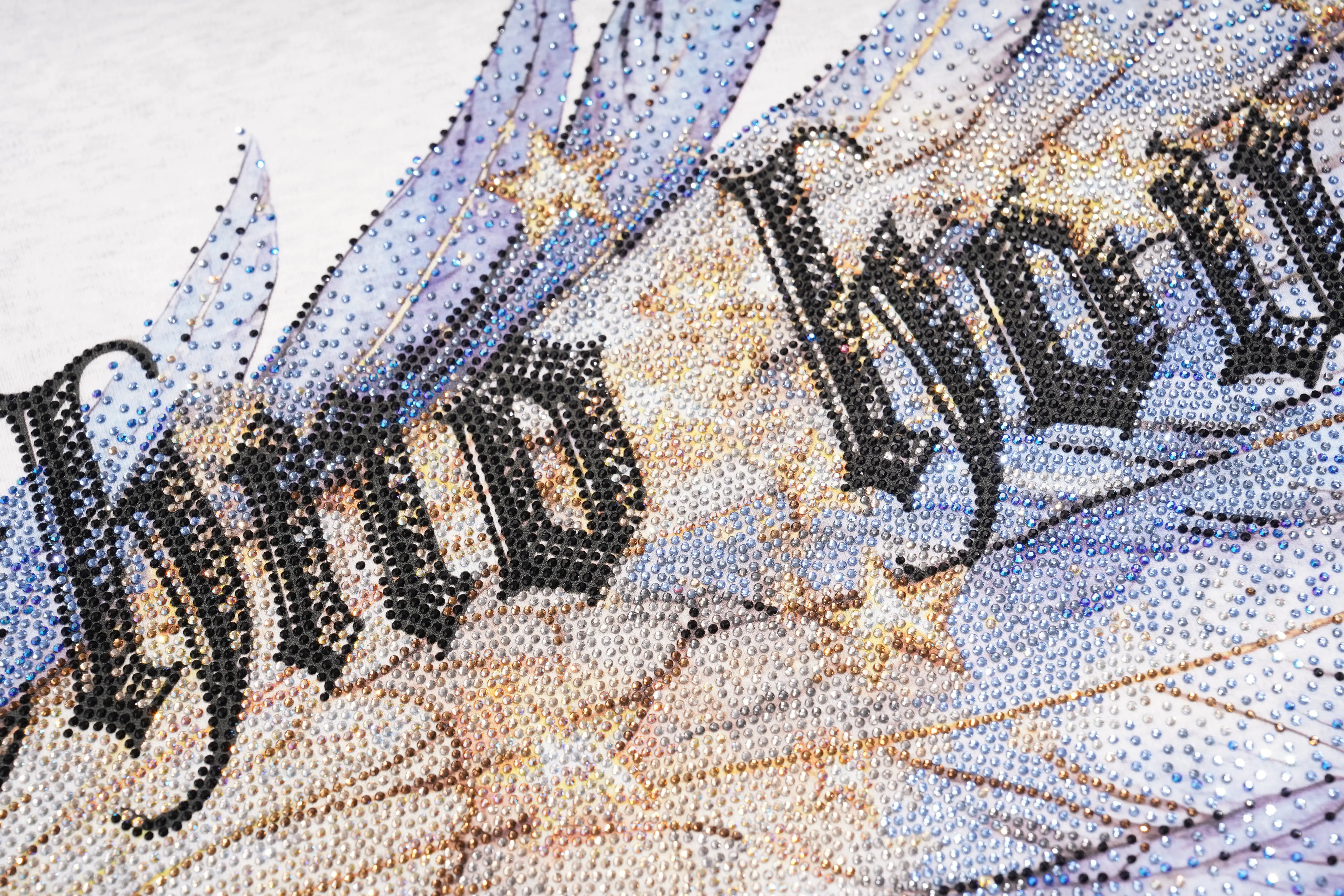 Close-up of a beaded wall art piece featuring the gothic-style lettering 'And How' on a wing-shaped design with blue, gold, and black beads on a white background. TCH Techno House Wing Graphic Oversized T-Shirt