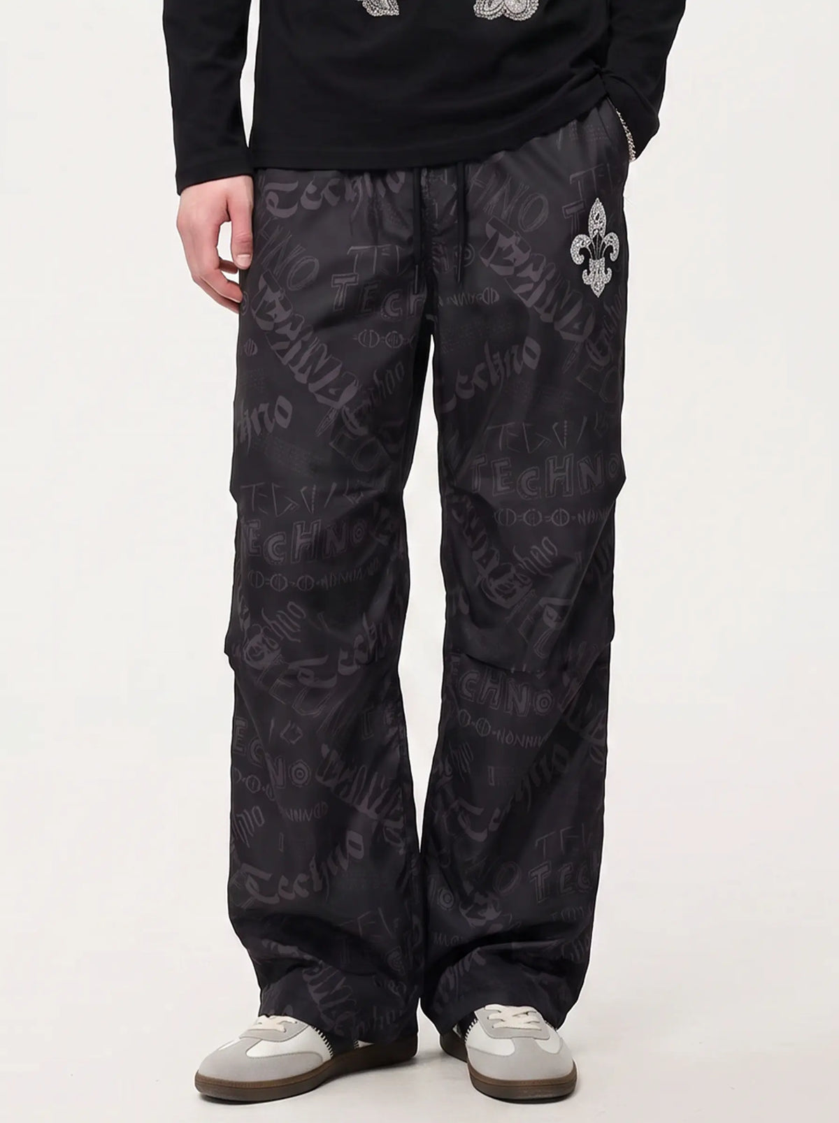 A person wearing black wide-leg pants with an allover tonal script pattern and a silver fleur-de-lis logo on the upper right thigh, paired with a black long-sleeve top and gray slip-on shoes, against a plain light background. TCH Techno Logo Wide-Leg Cargo Pants
